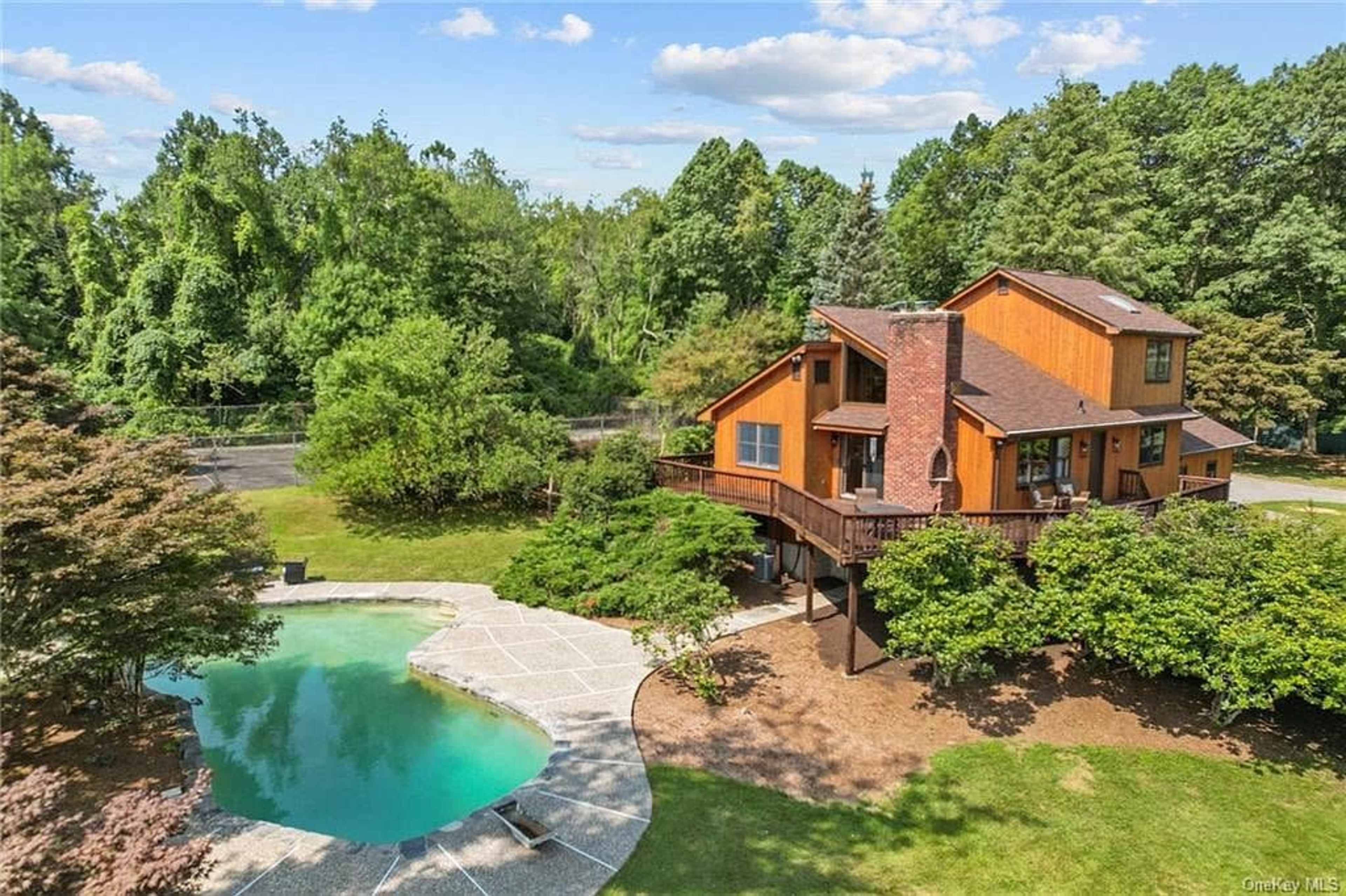 A two-story wooden house with large windows overlooks a curved swimming pool surrounded by landscaped greenery.
