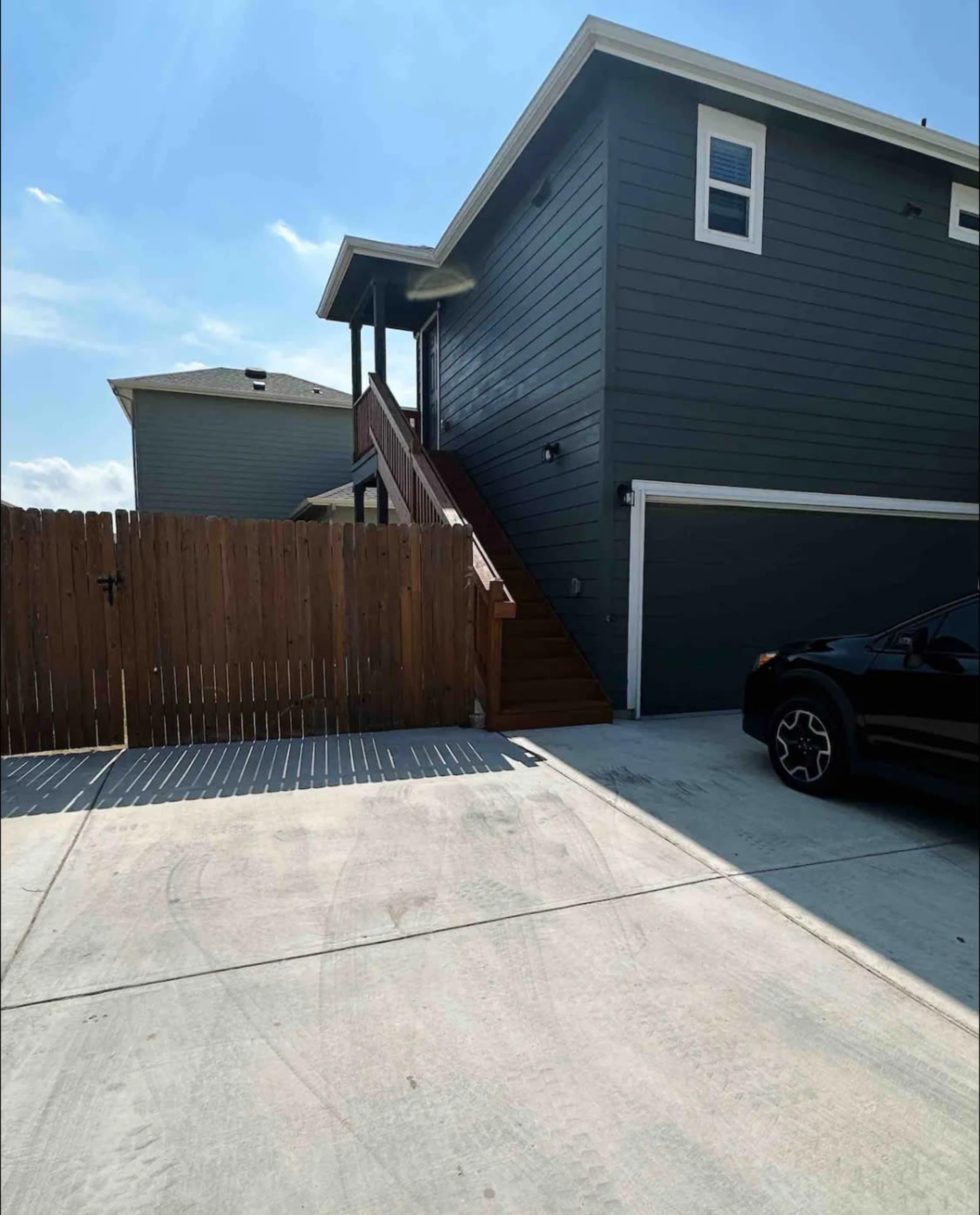 The image shows a two-story gray house with a wooden staircase leading to the second floor, next to a fenced yard and a parked black vehicle in the driveway.