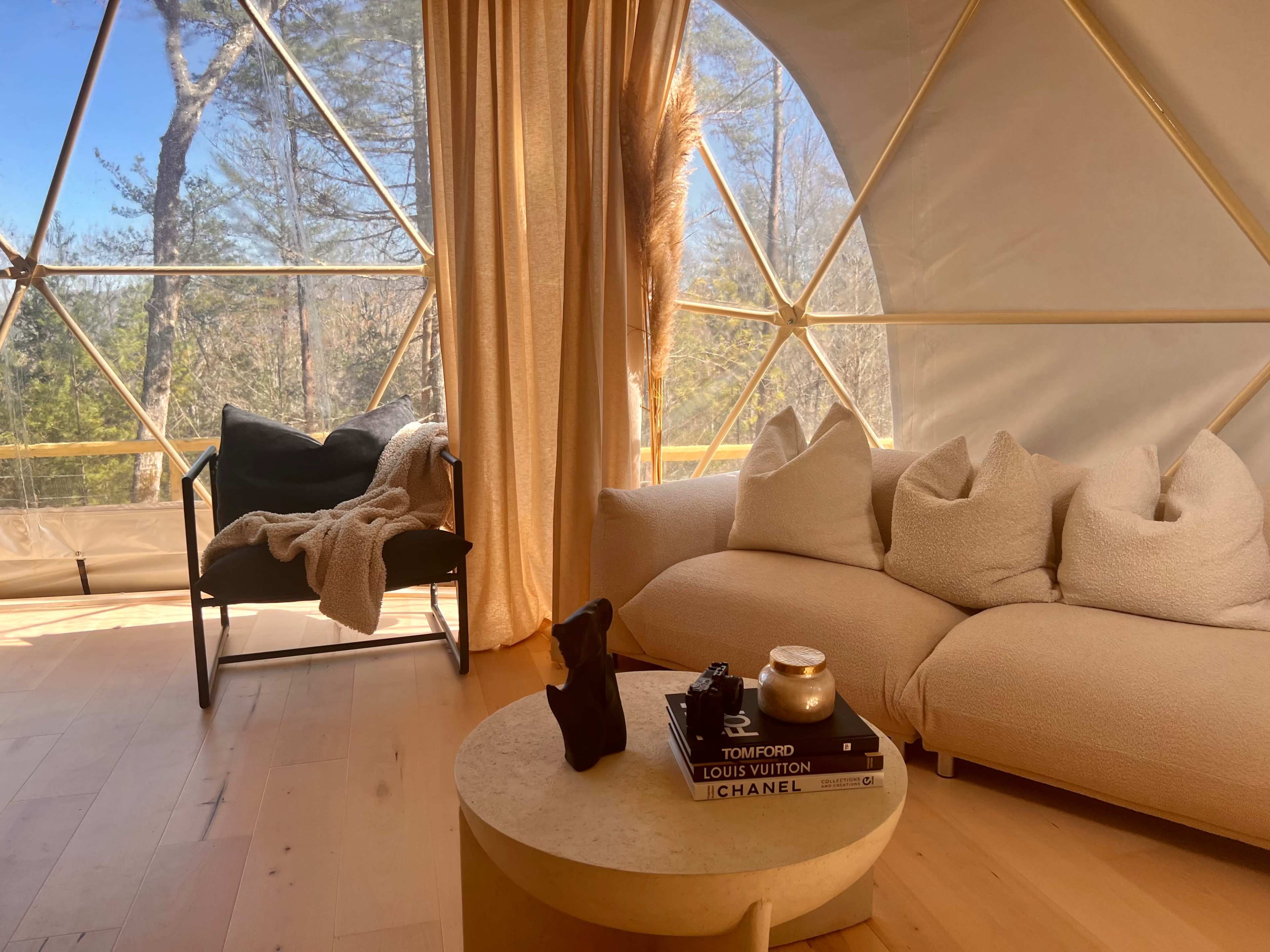 The interior of a geodesic dome features a light-colored sofa with accent pillows, a chair, a coffee table with decorative items, and large windows overlooking trees.