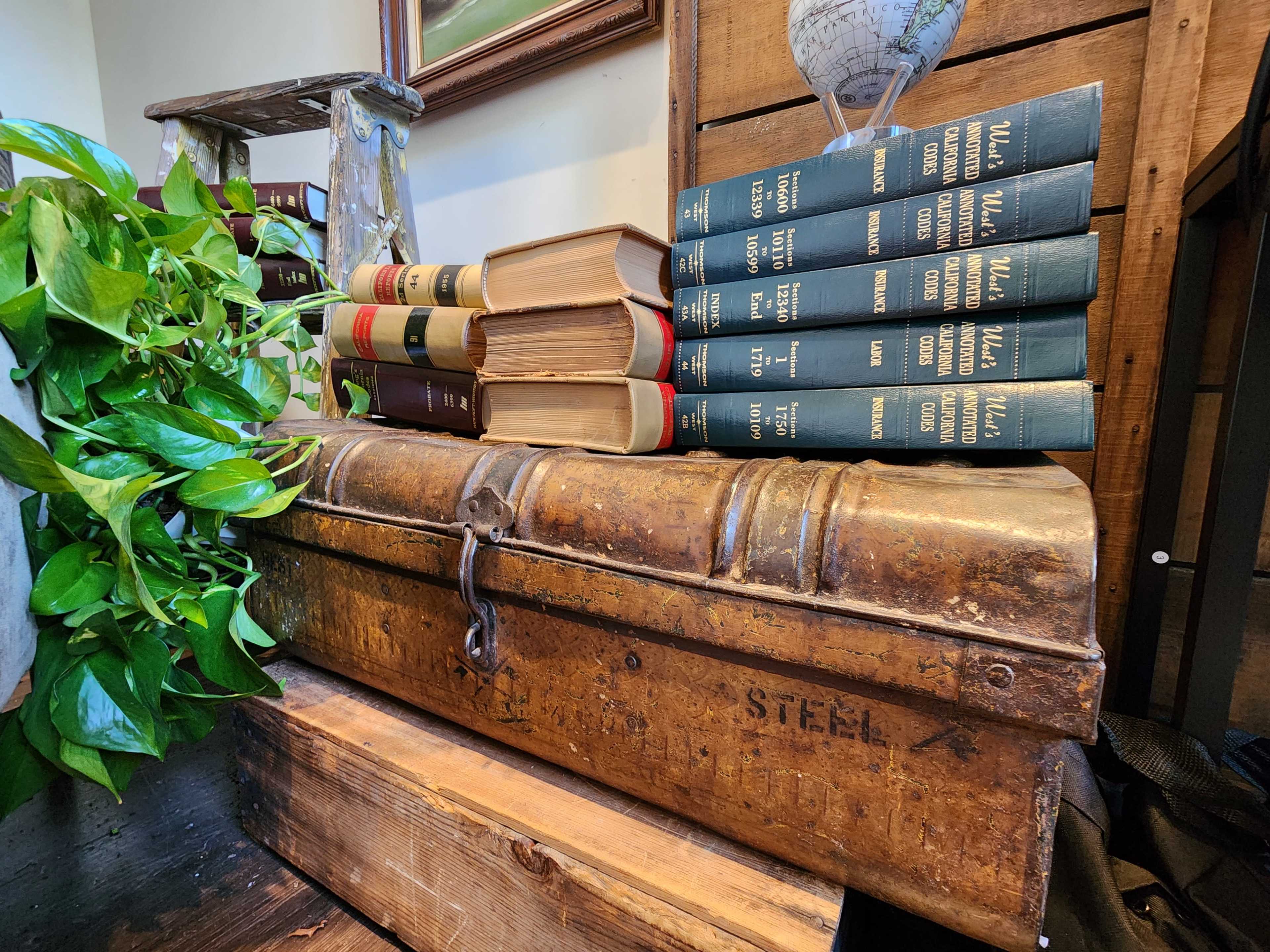 A stack of vintage books rests on an old trunk, with a green plant and a ladder in the background.