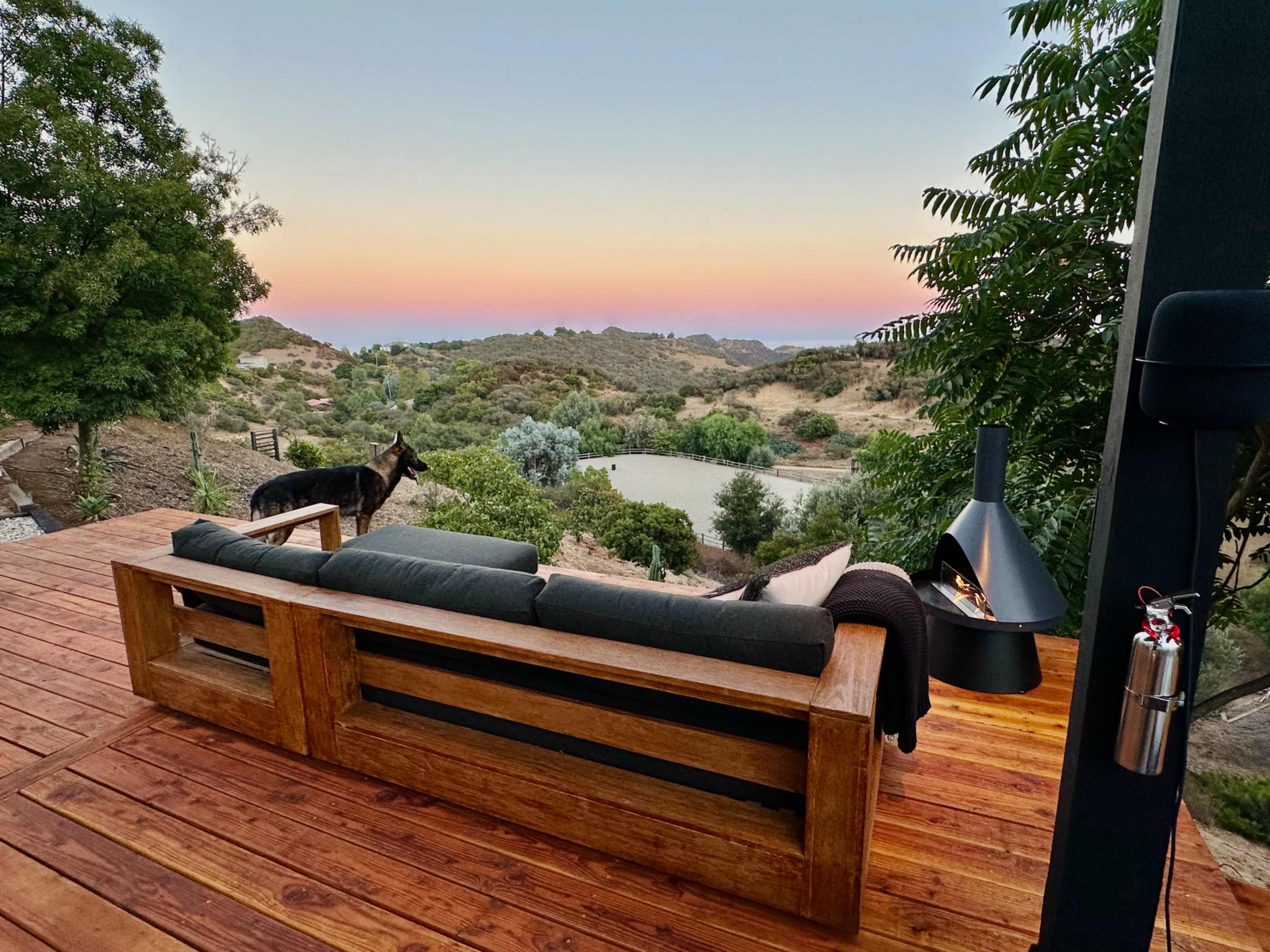 A wooden deck features a black sofa overlooking a grassy area and distant hills at sunset.