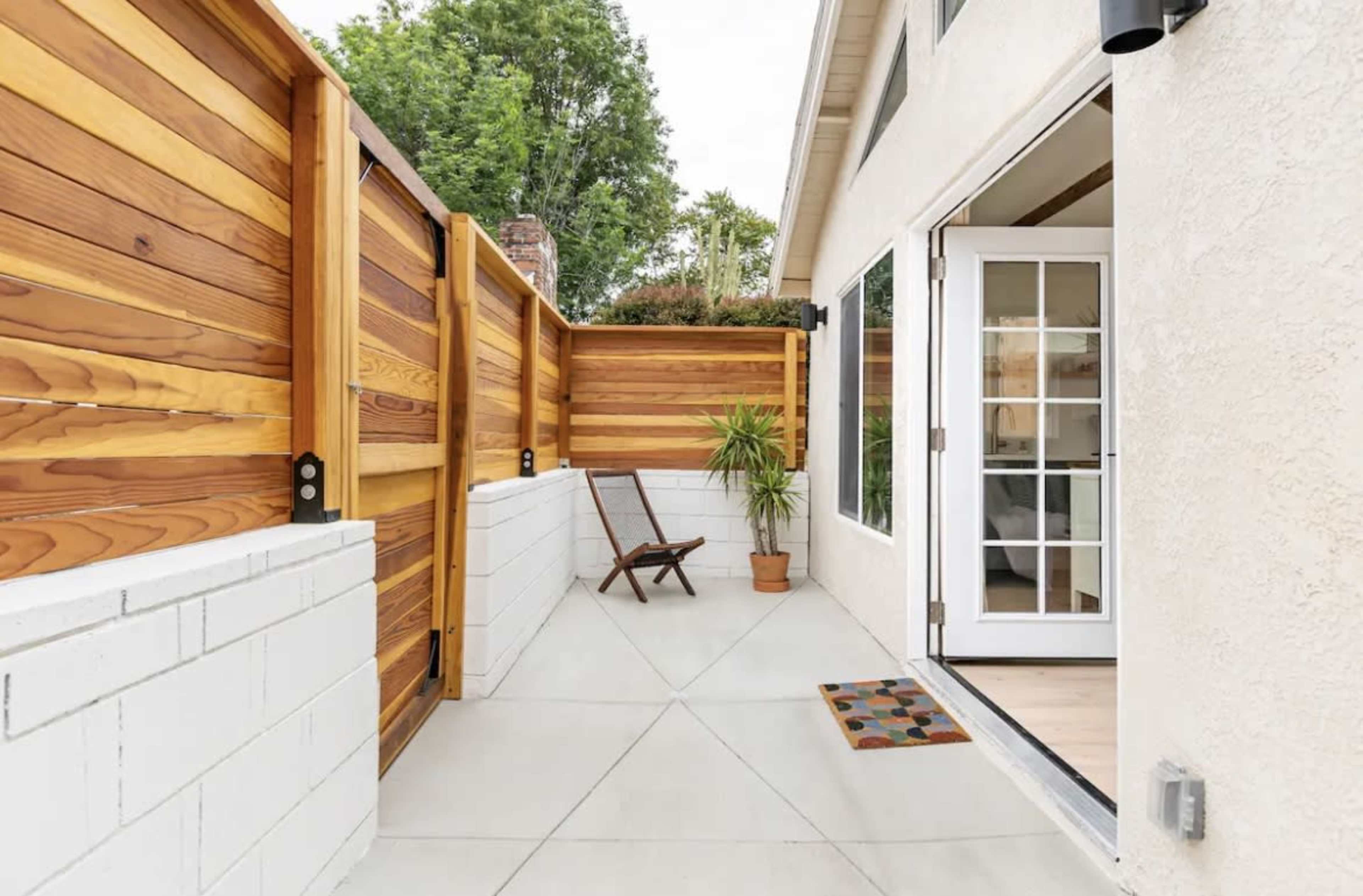 The image shows a small patio area enclosed by wooden fences, featuring a potted plant and a folding chair next to a door with French-style windows.