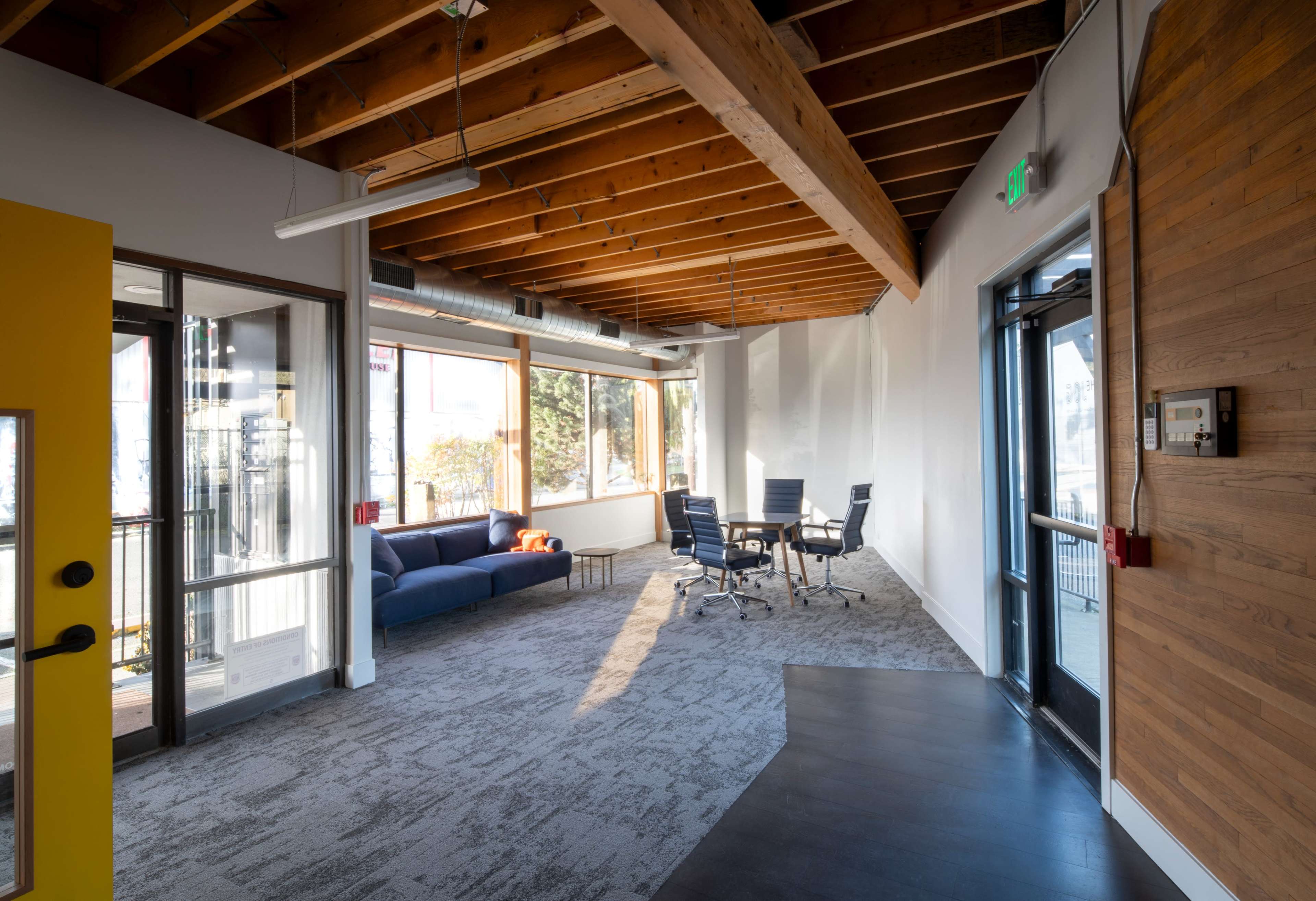 A modern office space with a yellow door, a blue couch, a conference table surrounded by chairs, and exposed wooden beams on the ceiling.