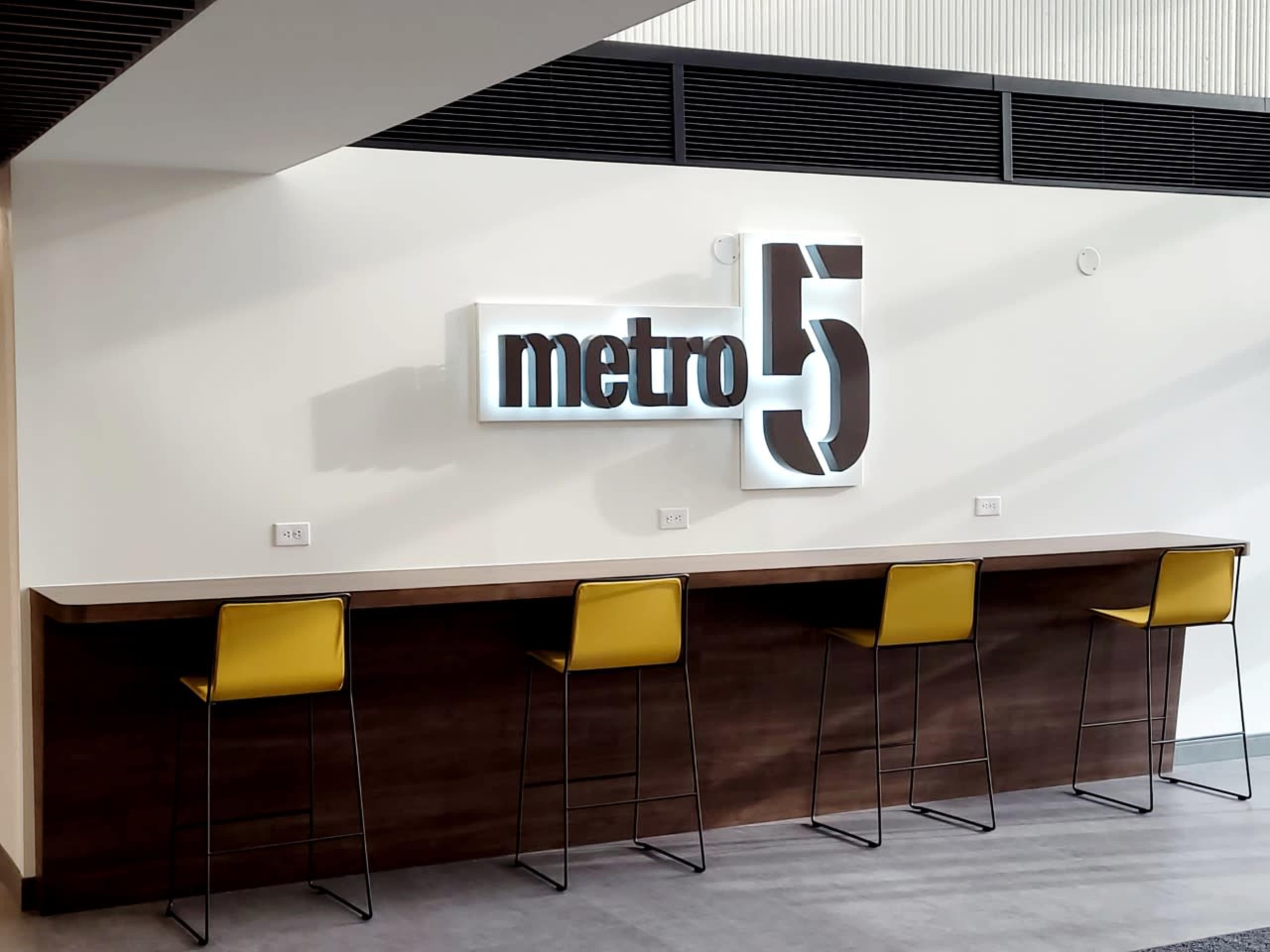 A modern bar area featuring a long wooden counter with four yellow chairs and a large illuminated sign reading "metro 5" on the wall behind it.