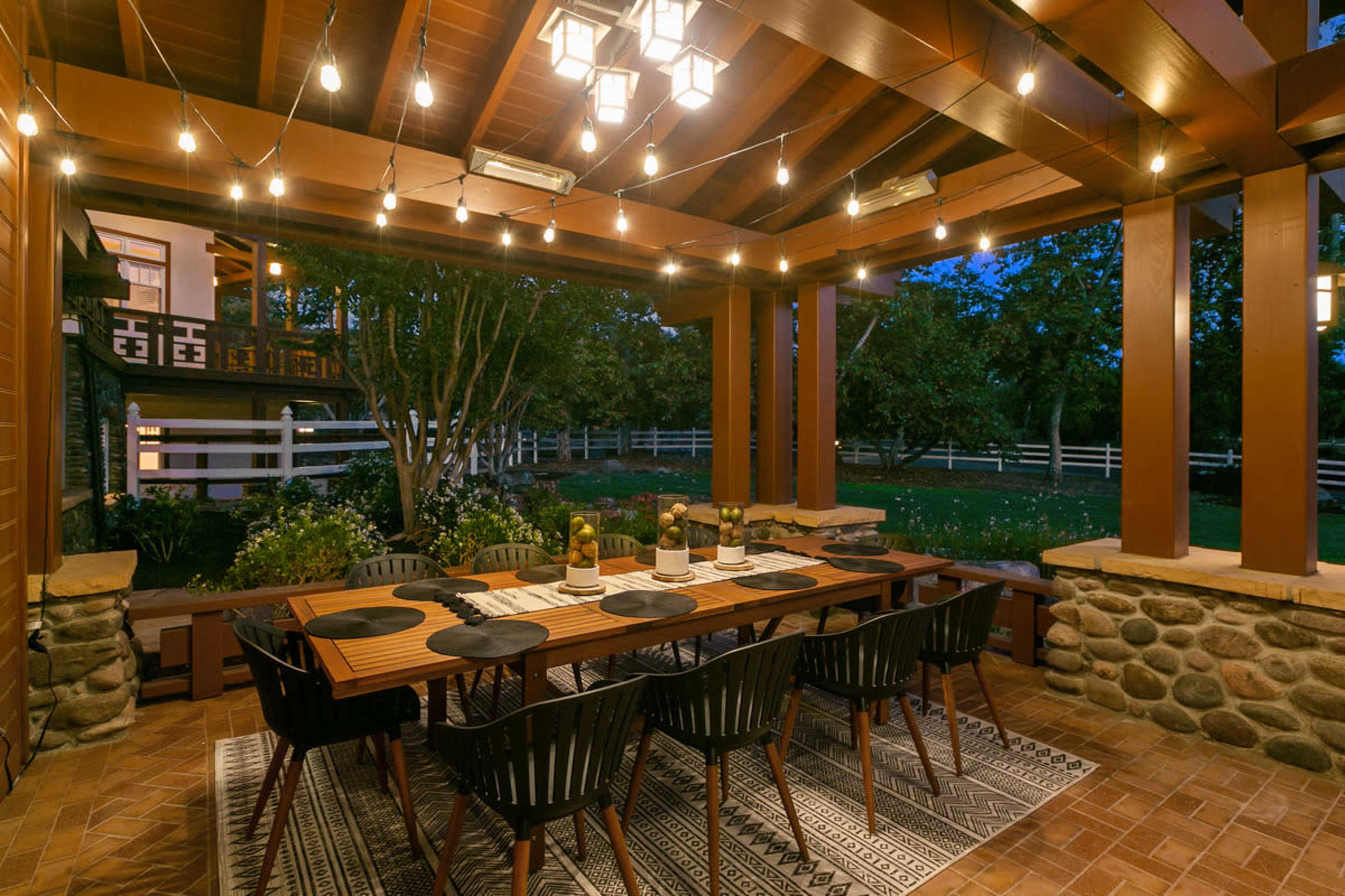A spacious outdoor dining area features a long wooden table set for dinner, surrounded by black chairs and illuminated by string lights overhead.