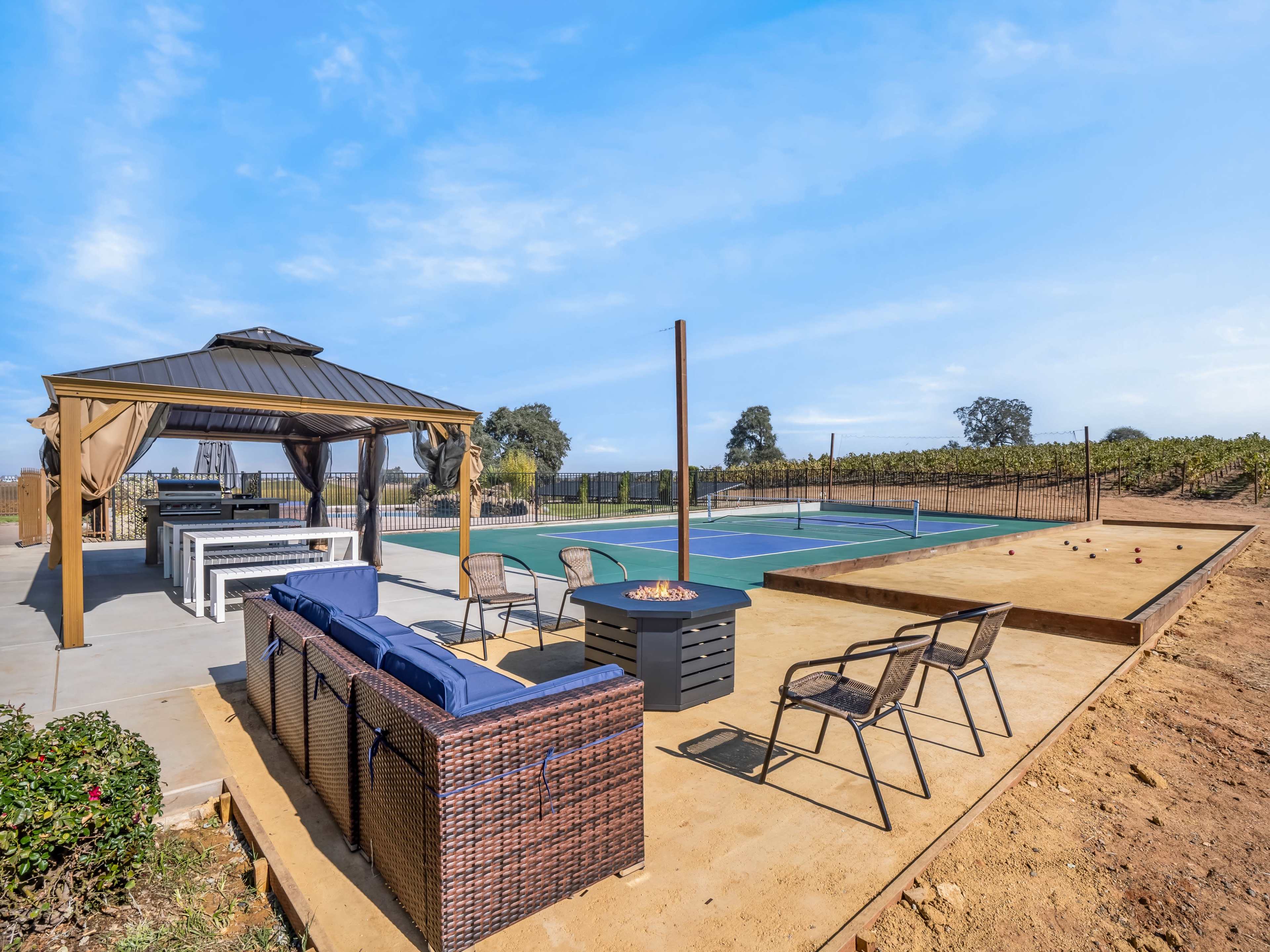 A patio area with seating and a fire pit beside a tennis court, surrounded by vineyards.
