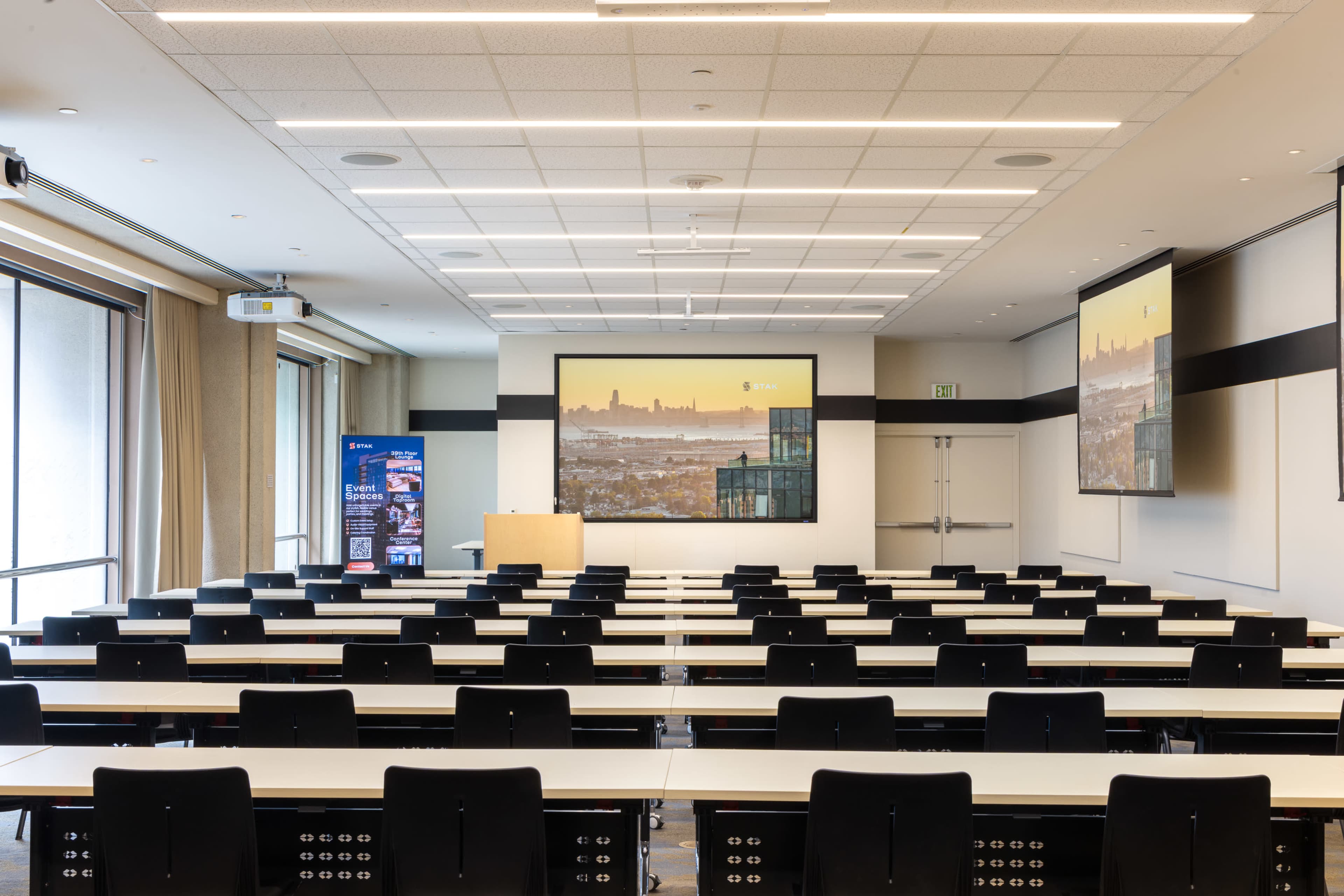 The image shows a modern conference room with rows of tables and chairs, large screens displaying city views, and large windows allowing natural light.