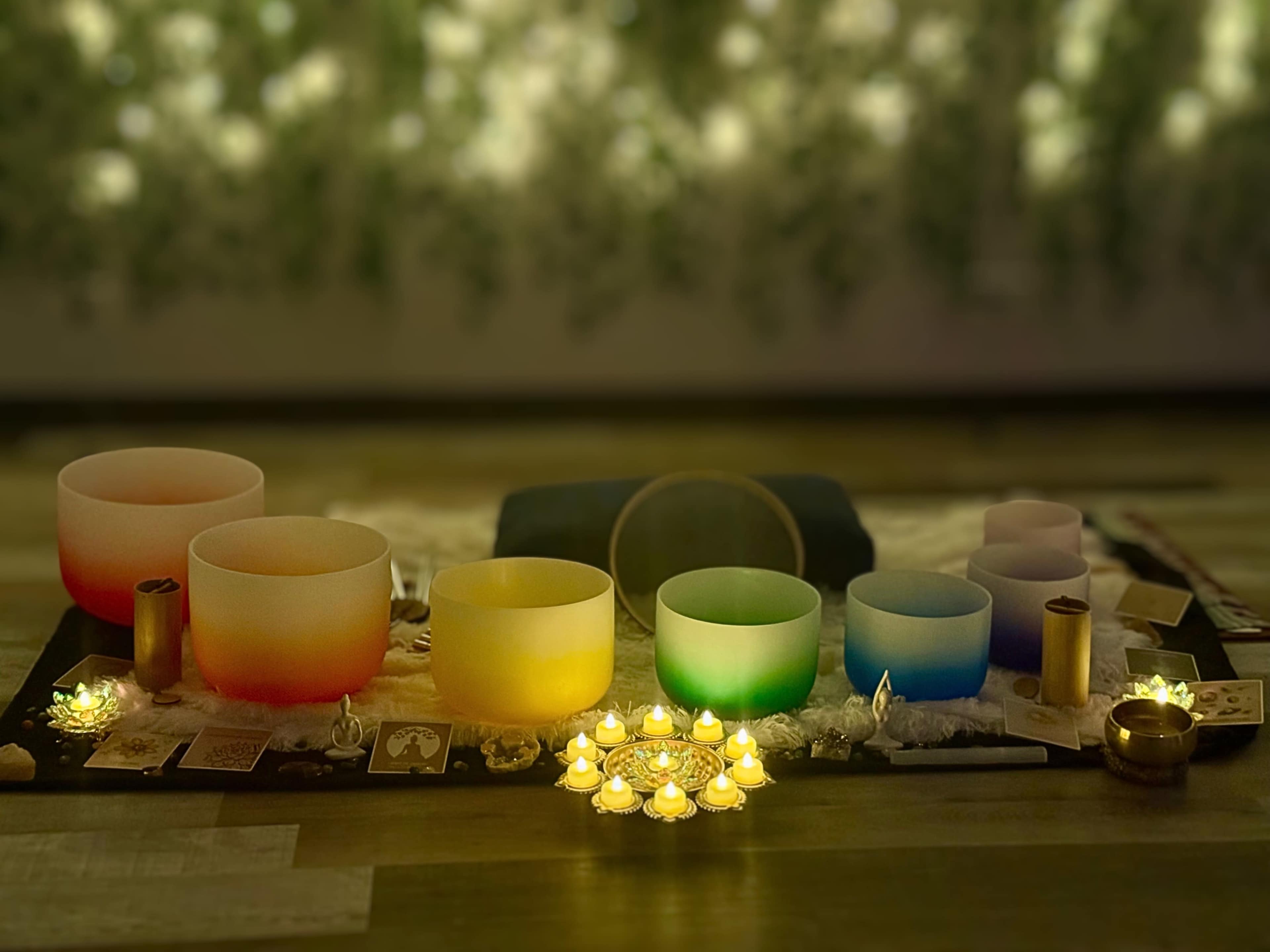 The image shows a serene sound healing setup featuring colorful crystal singing bowls, lit candles arranged in a circle, and various spiritual items on a textured surface.