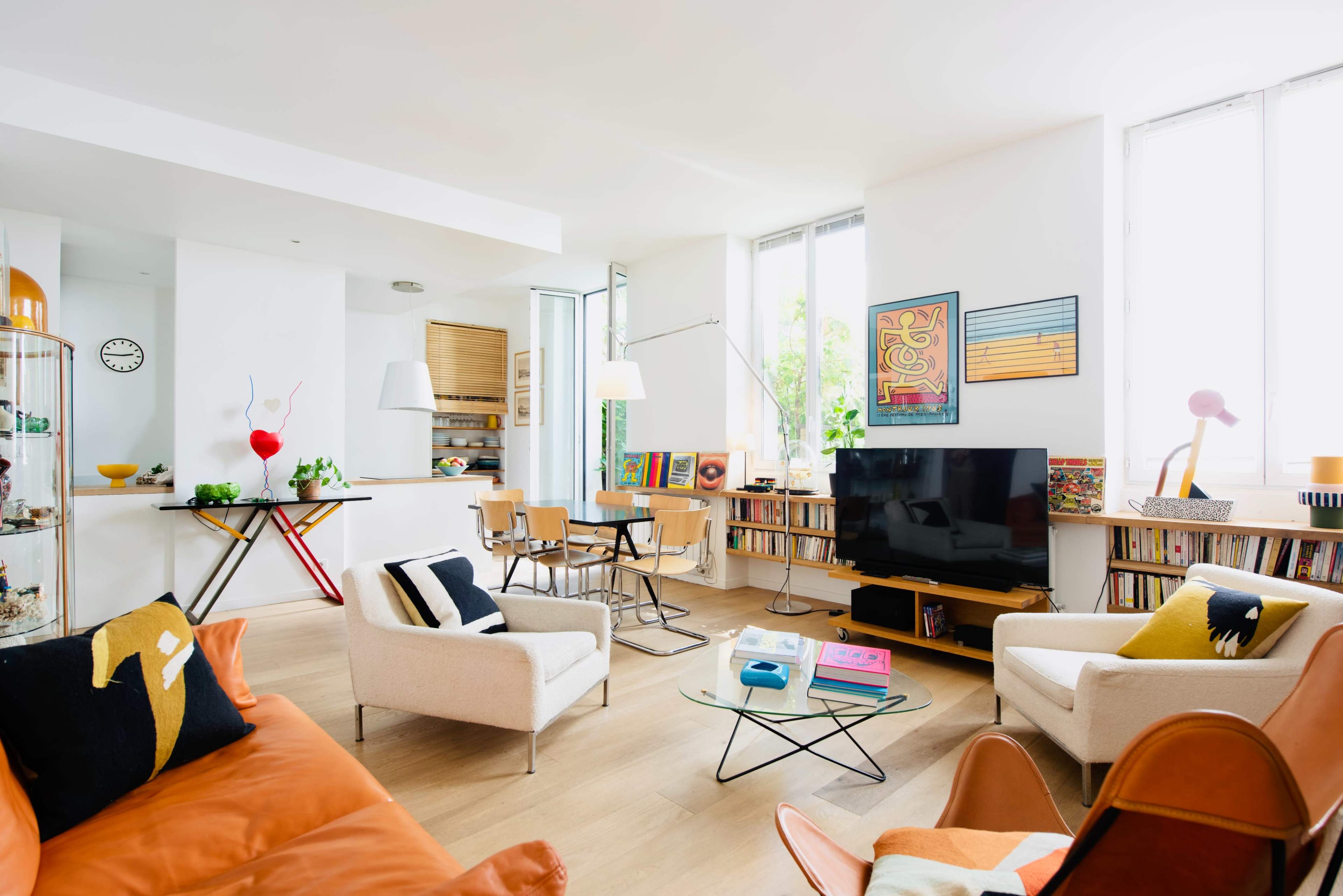 The image shows a modern living room with a mix of seating options, a television, bookshelves, a dining area, and large windows allowing natural light.