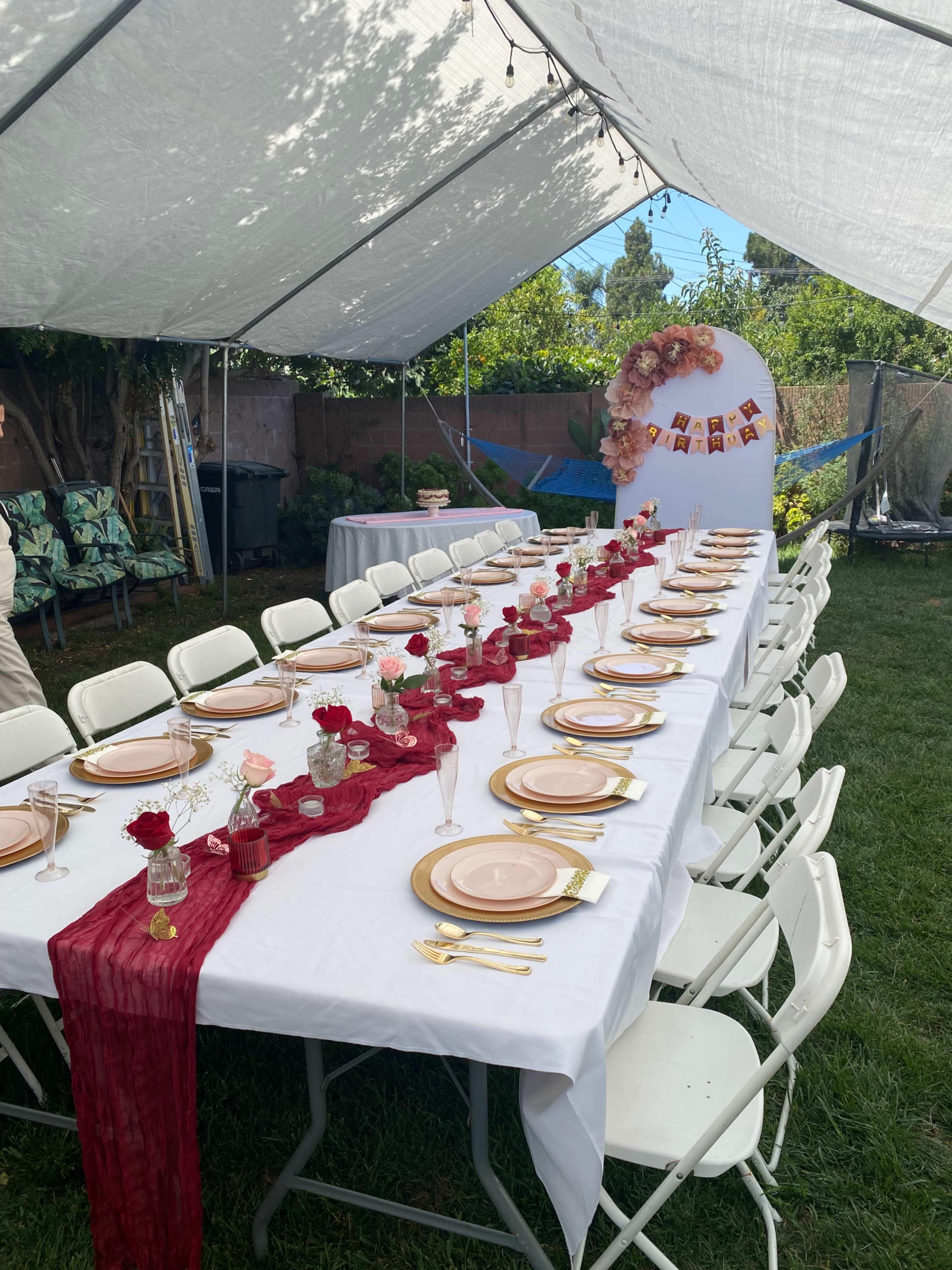 A long table is set up under a tent in a backyard, decorated with pink plates, red table runners, and floral centerpieces.