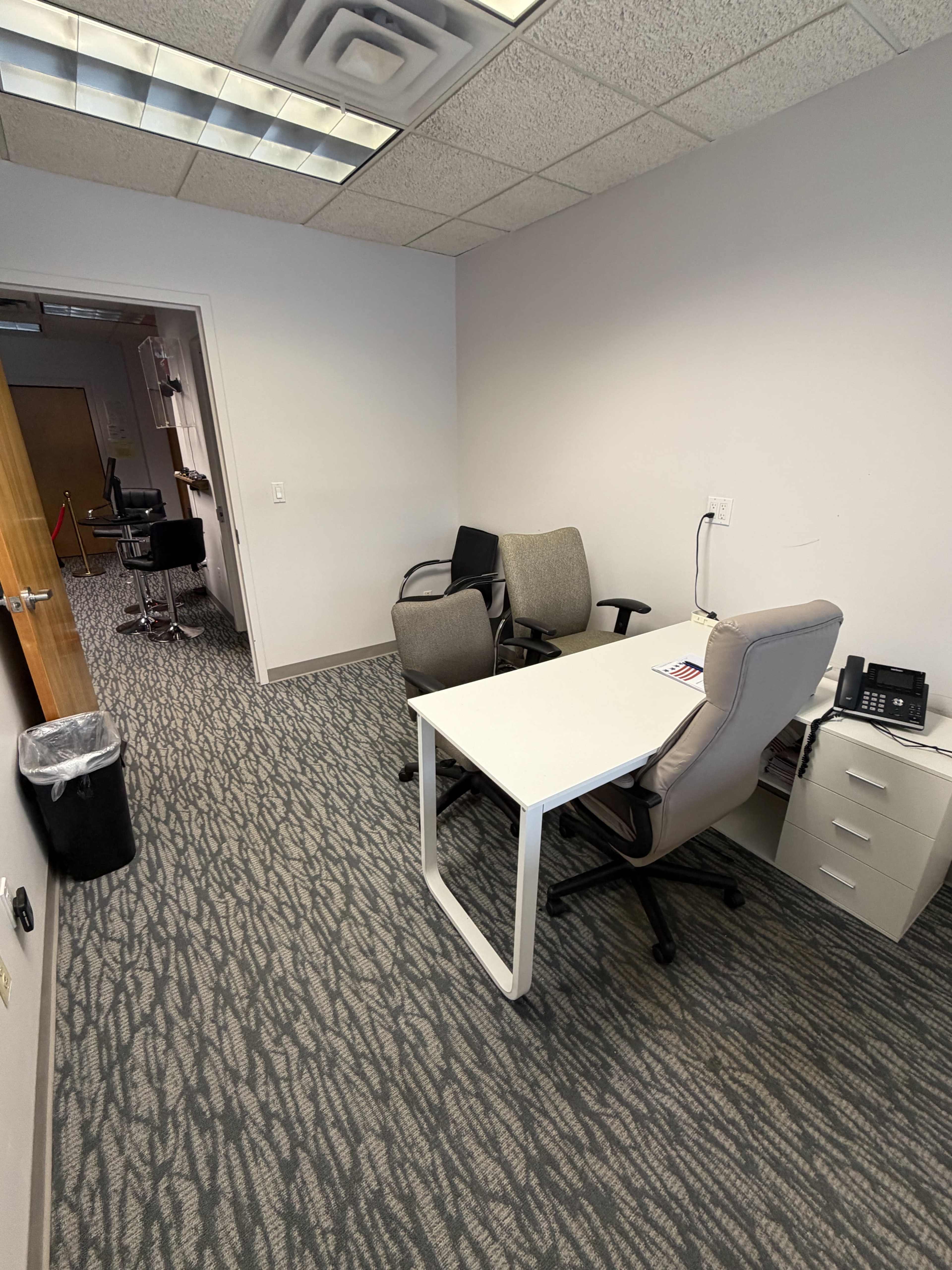 The image shows a sparsely furnished office with a white desk, a chair, and a filing cabinet, along with a trash can in the corner.