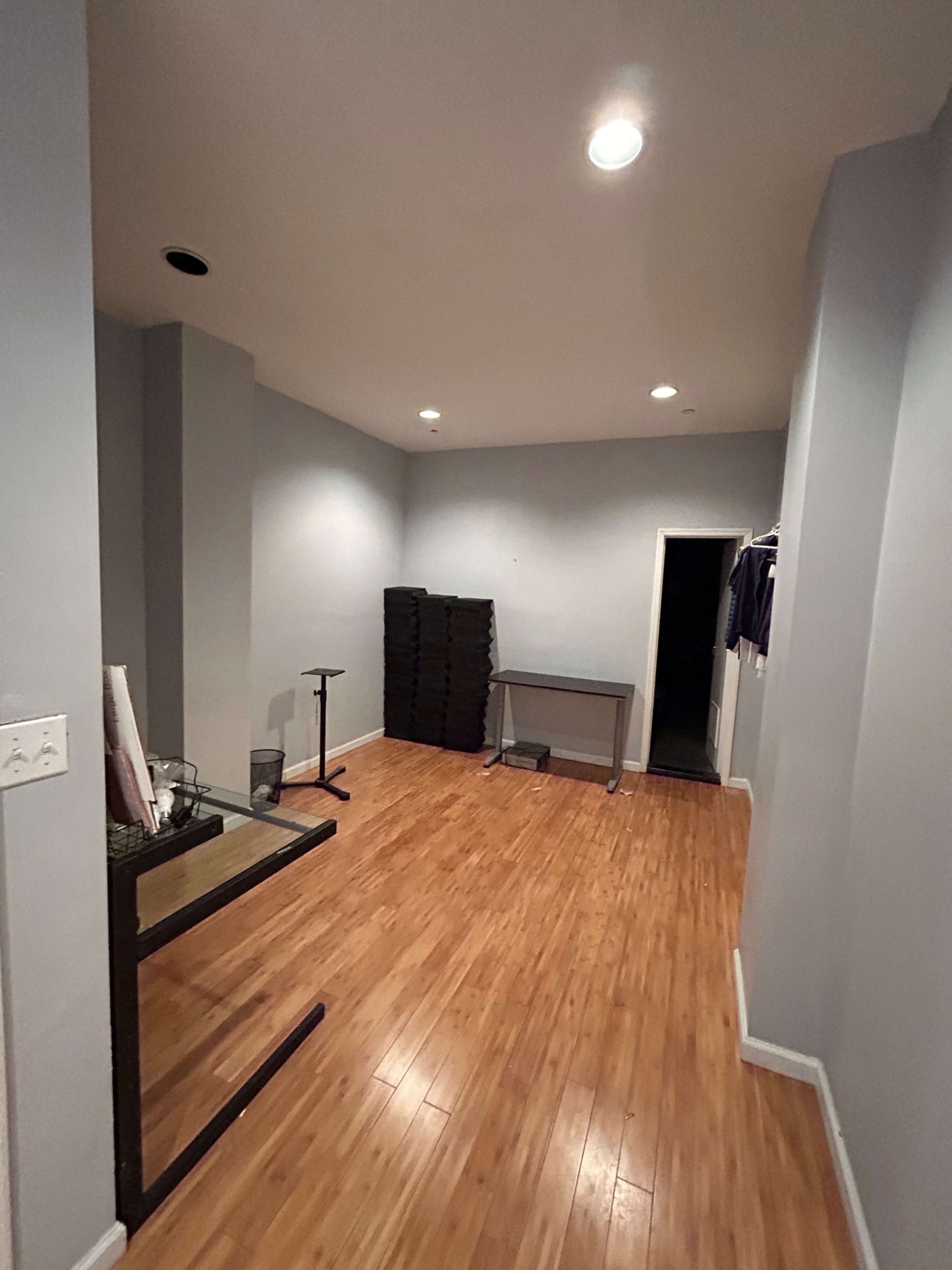 The image shows a vacant room with wooden flooring, pale gray walls, a glass table, and a stack of boxes against one wall.