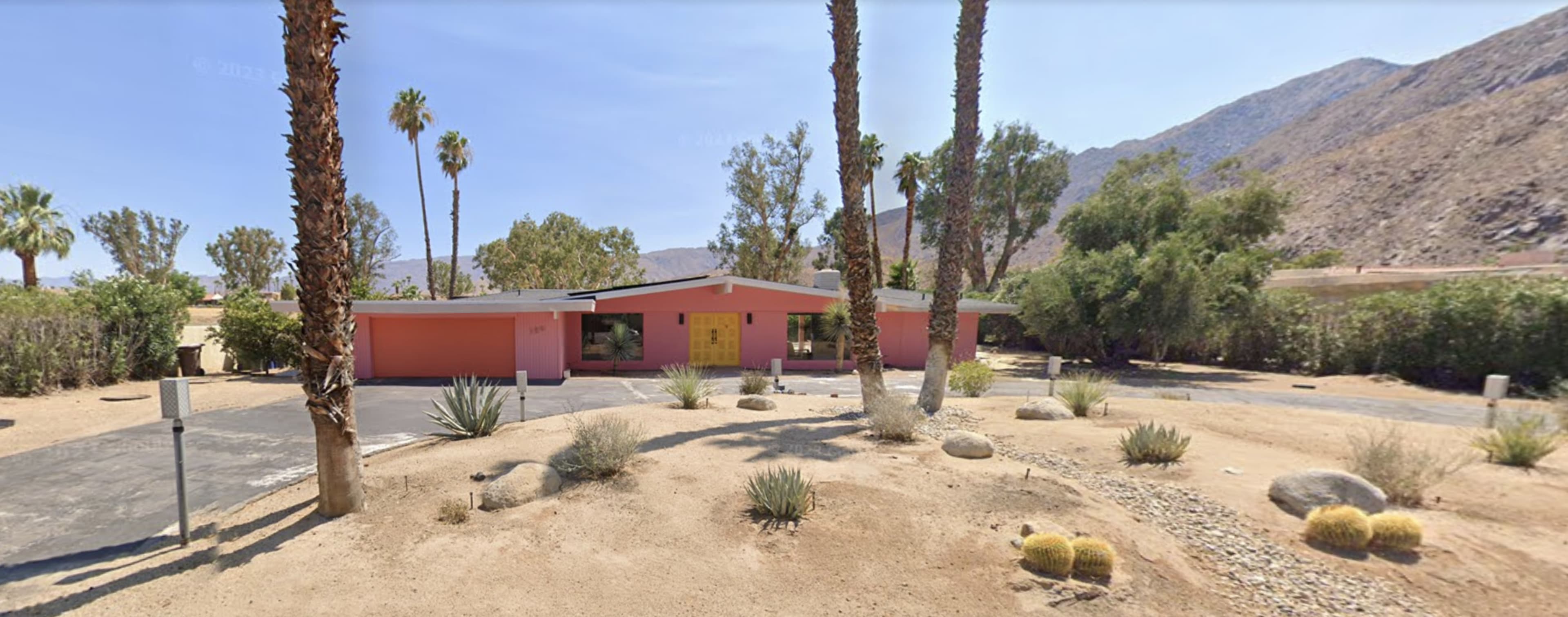 A single-story pink house with a yellow door sits among palm trees and desert landscaping against a mountainous backdrop.