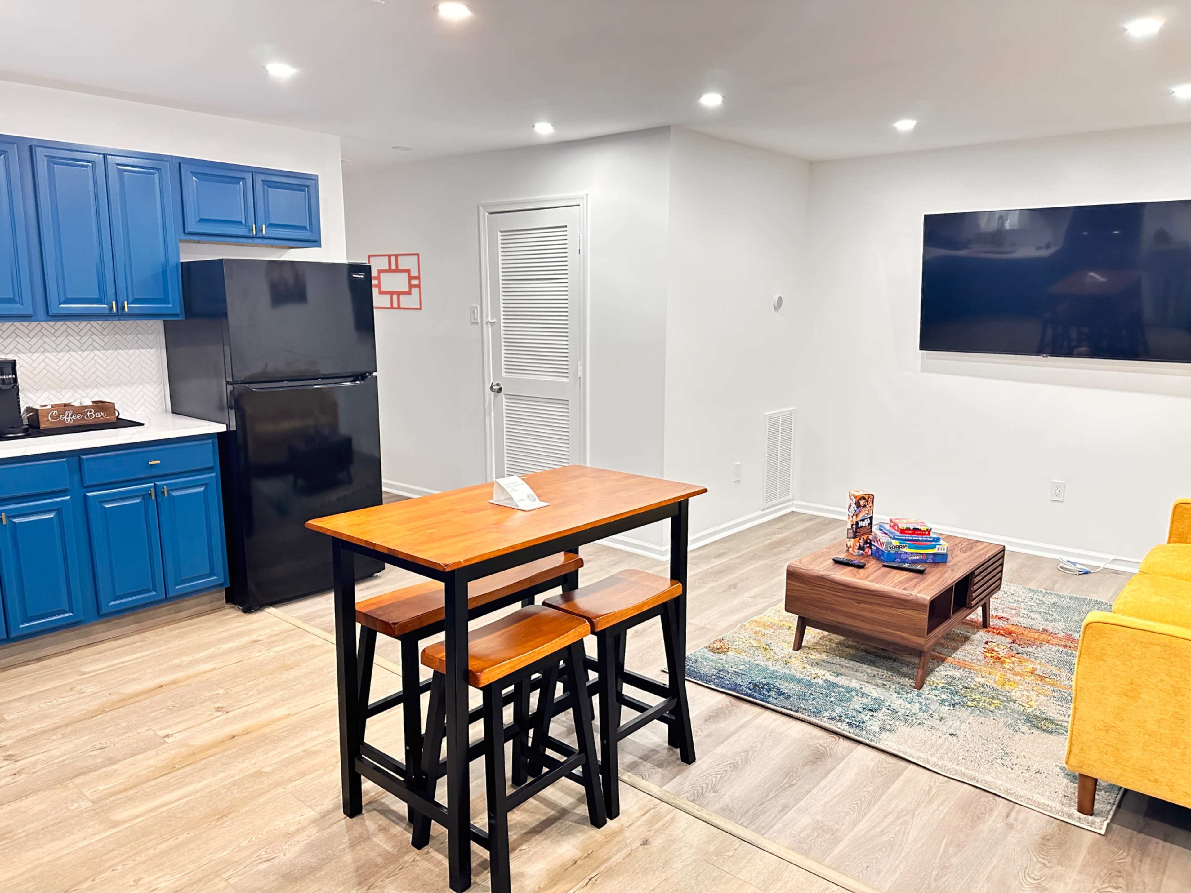 The image shows a modern living space featuring a kitchen area with blue cabinets and a black refrigerator, a dining table with stools, a yellow couch, and a wall-mounted television.