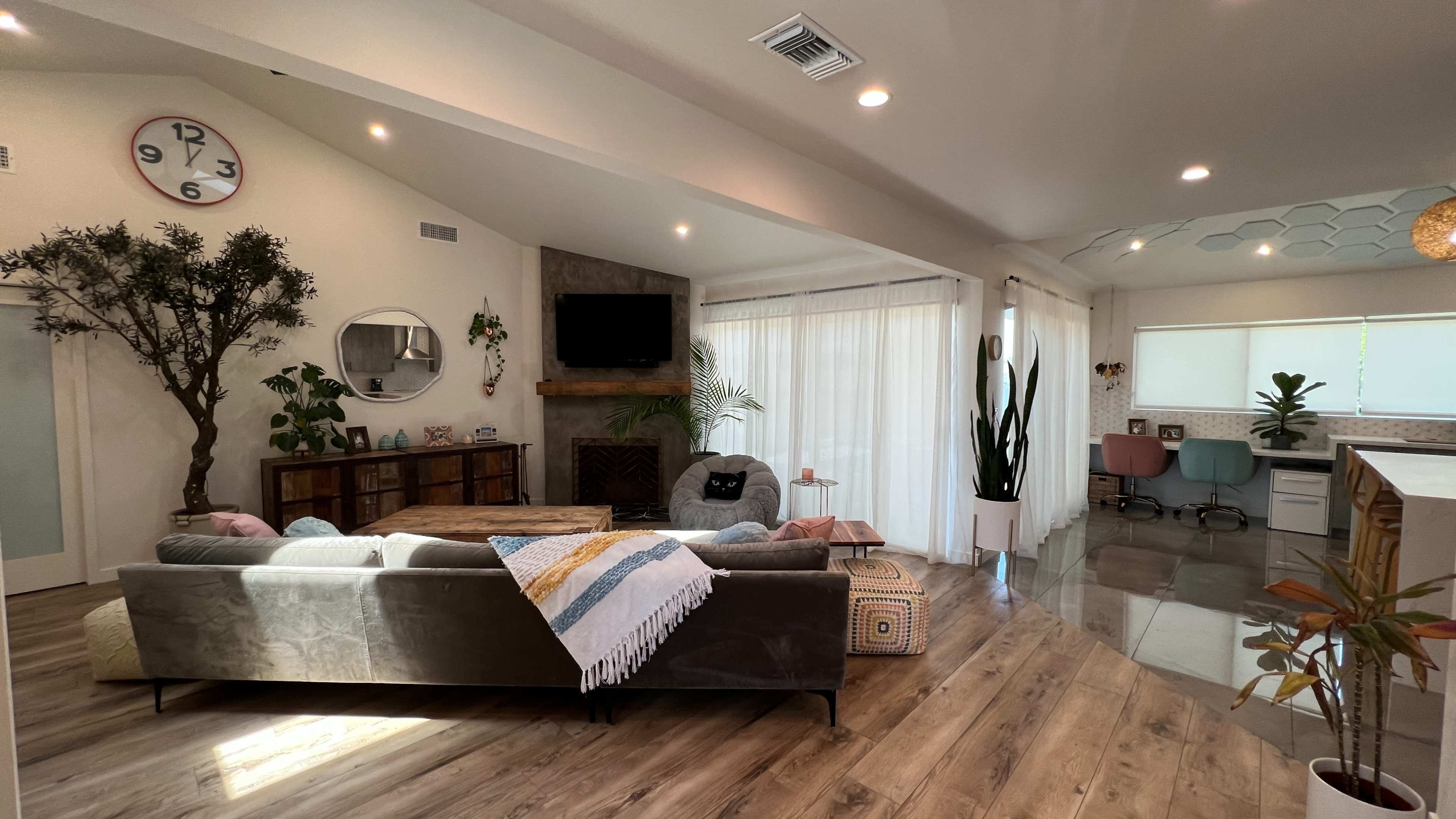 A modern living room with a large sectional sofa, a coffee table, a decorative wall clock, and large windows with sheer curtains, leading into a kitchen area with two pastel-colored chairs.