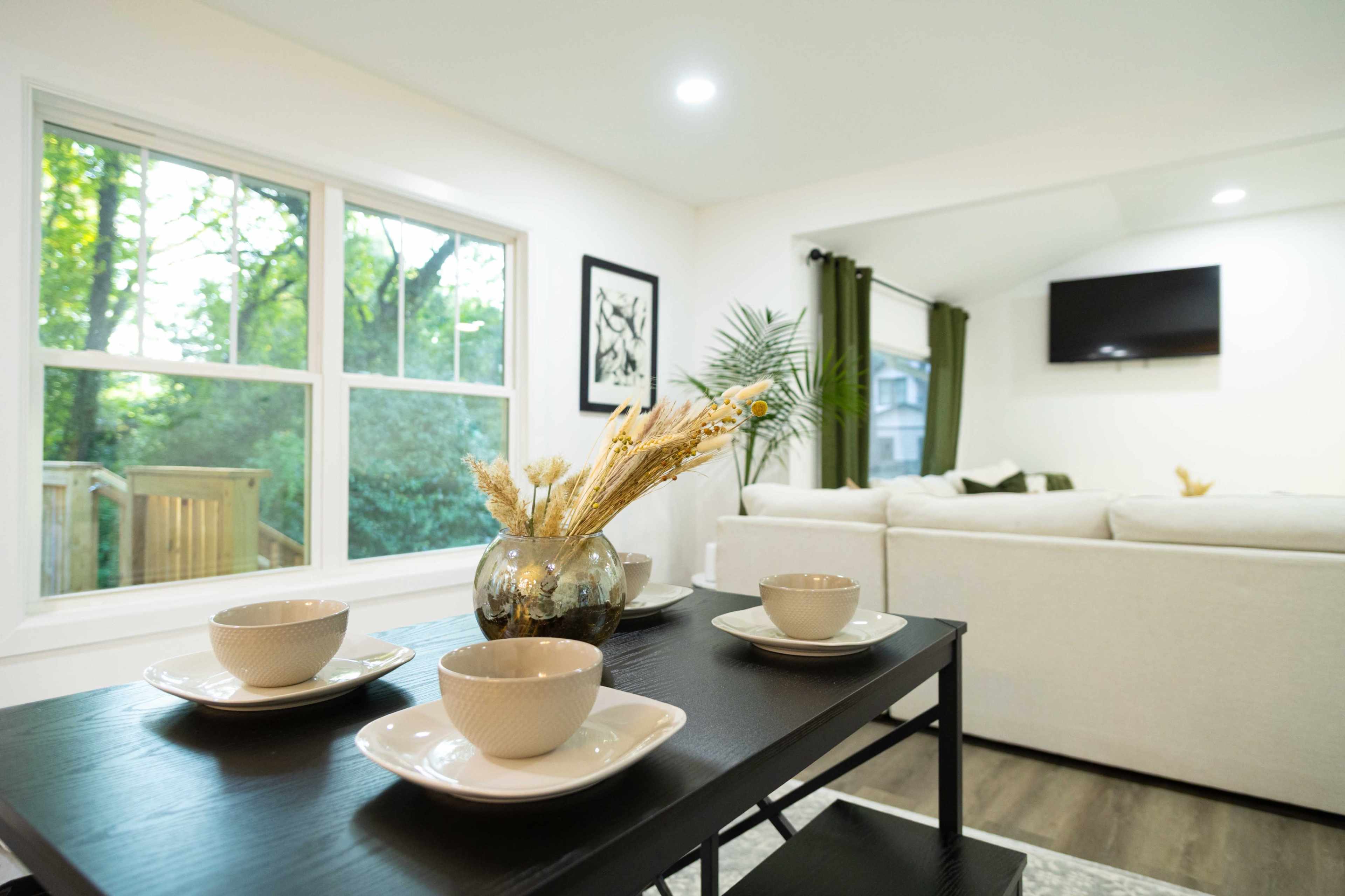 A modern living room features a black coffee table set with plates and bowls, surrounded by large windows showcasing greenery outside.