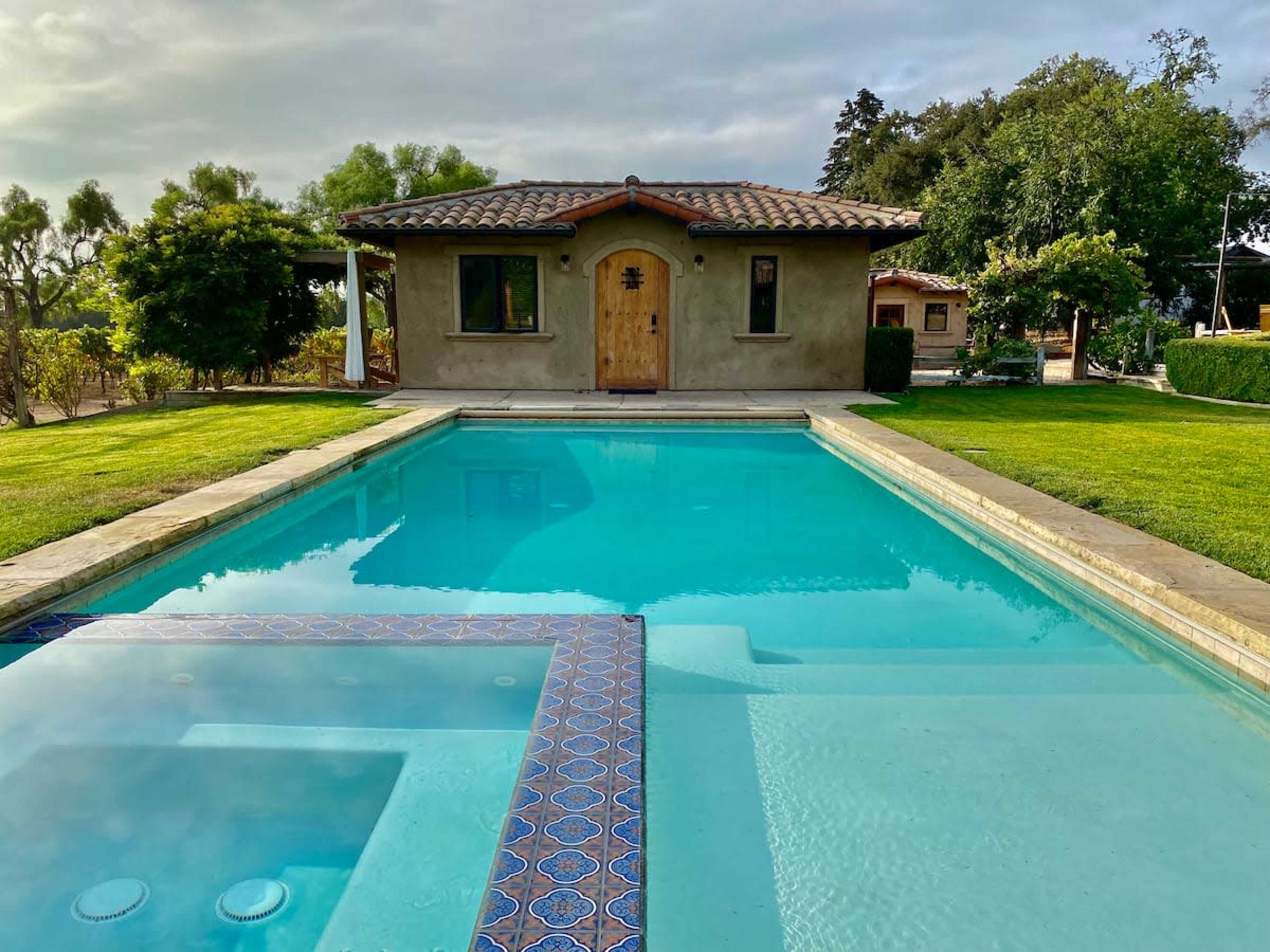 A clear swimming pool reflects a small house with a wooden door, surrounded by green grass and trees.