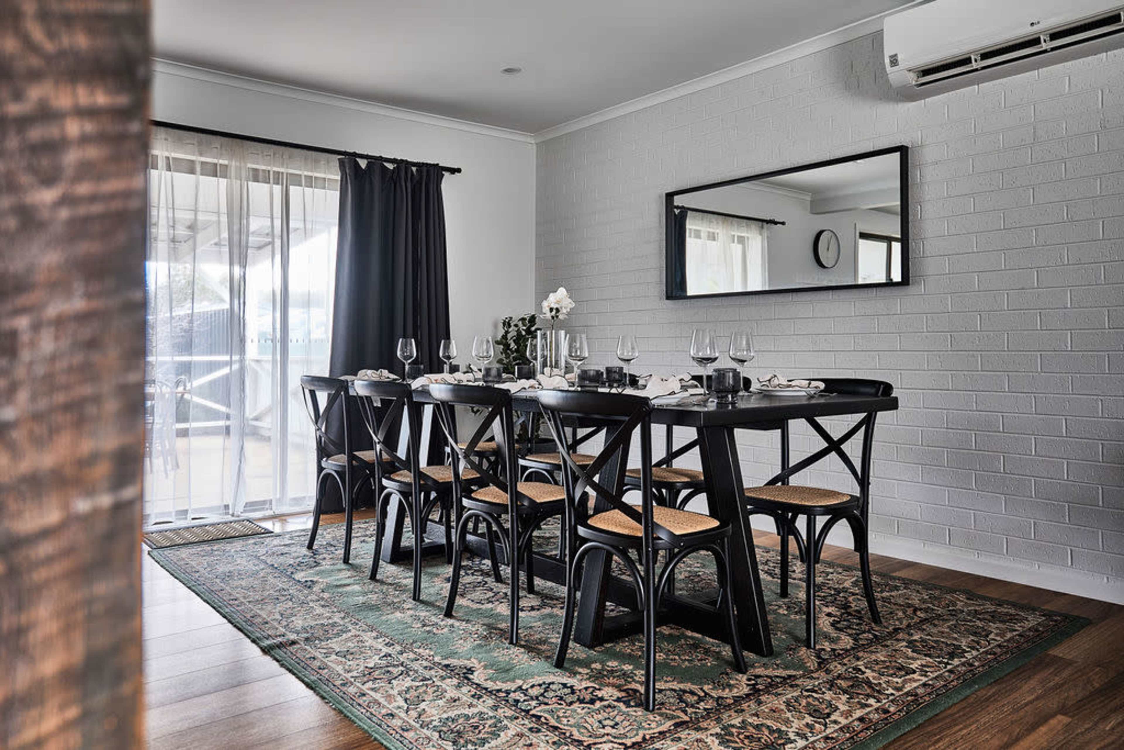 A dining room features a large black table with eight chairs, set with glasses and a centerpiece, alongside a patterned rug and a large mirror on the wall.