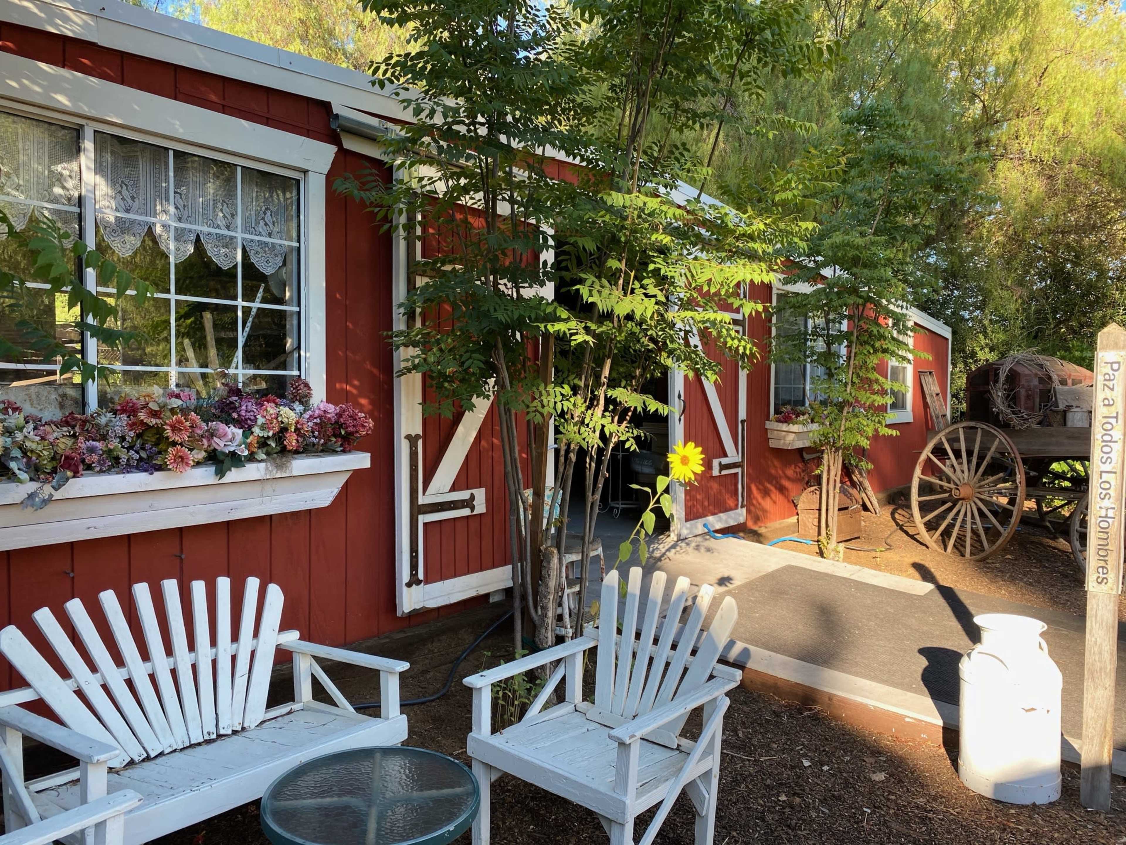 A red wooden building with white trim, featuring a front porch with two white chairs and a flower box, alongside a large wheel and a white container.