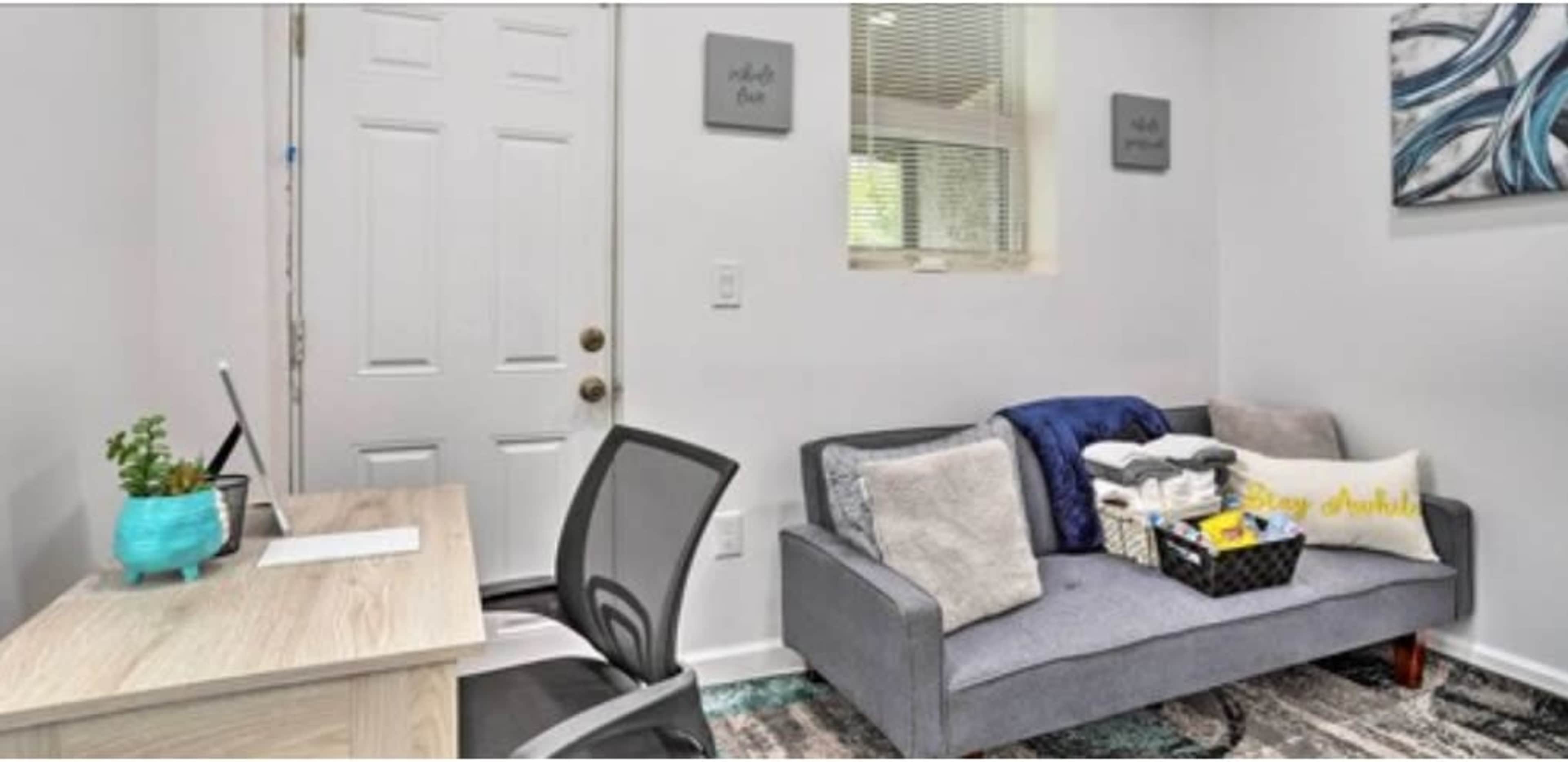 The image shows a small office space with a desk, chair, and a gray couch adorned with blankets and a basket of items against a white wall.