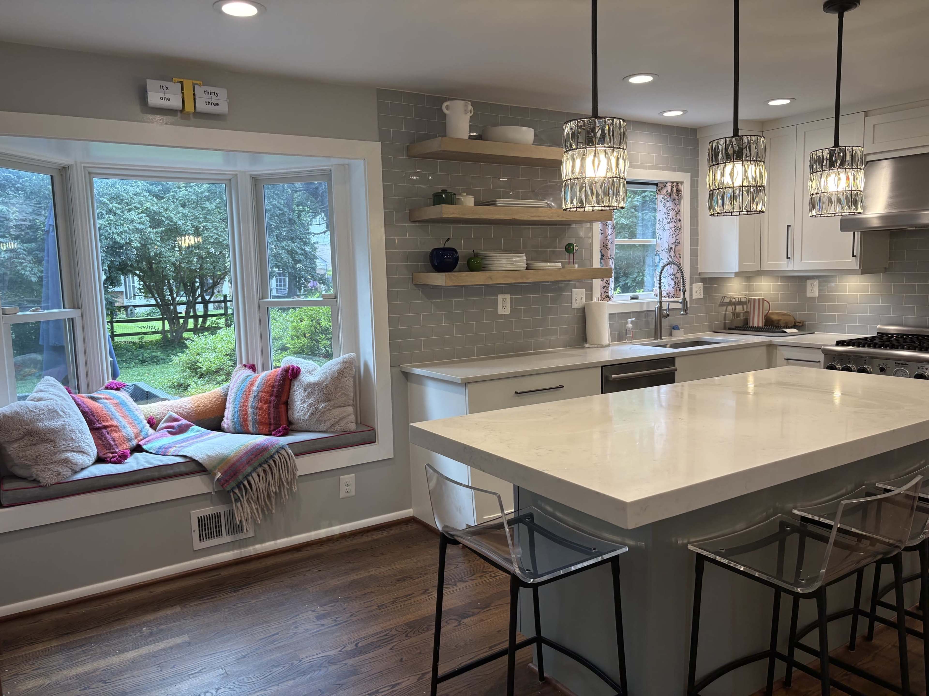 The image shows a modern kitchen featuring a large island with bar stools, open shelves, and a cozy window seat adorned with cushions.
