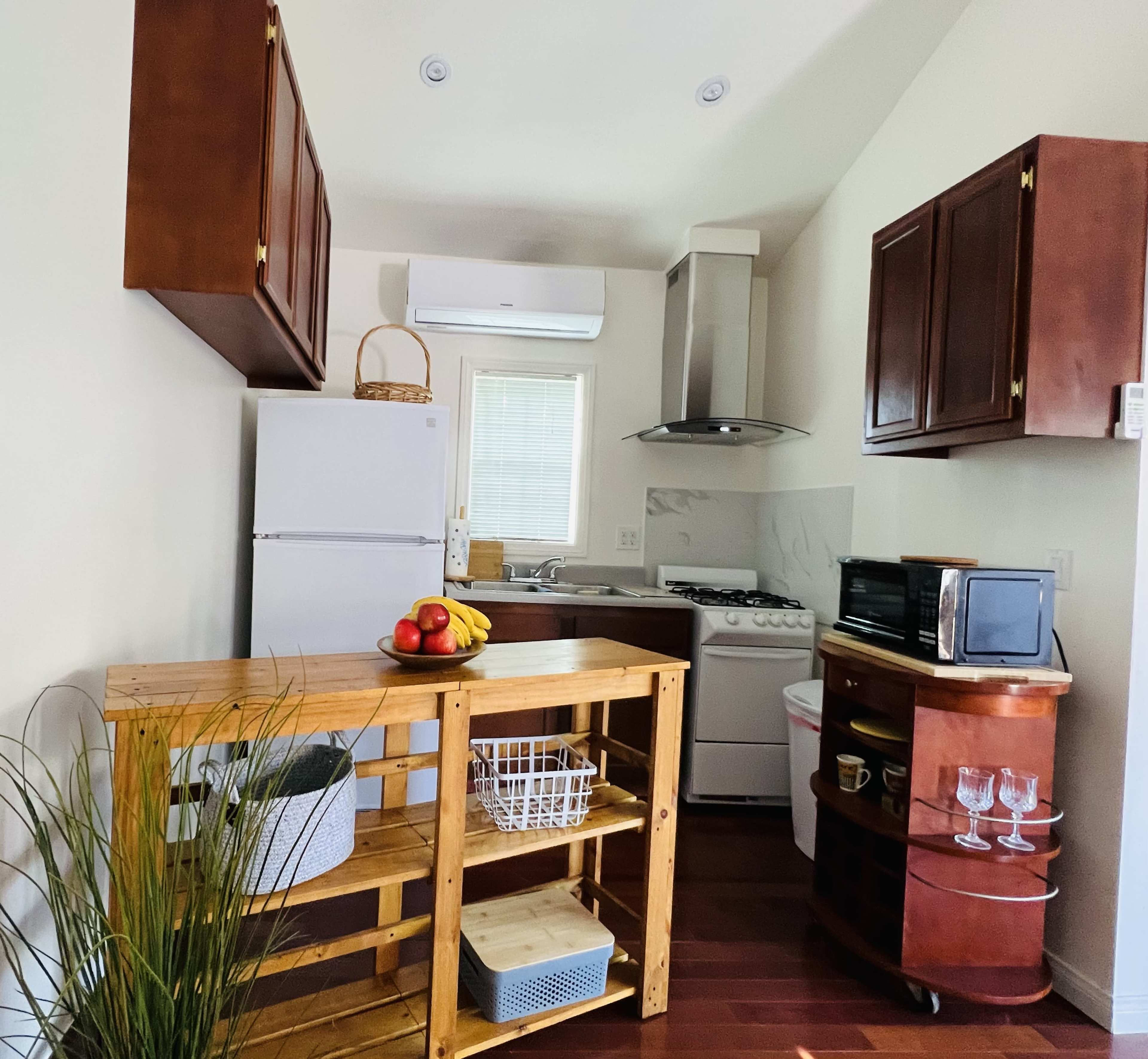 A small kitchen features brown wooden cabinets, a white refrigerator, a gas stove, and a wooden cart with fruit and dishware.