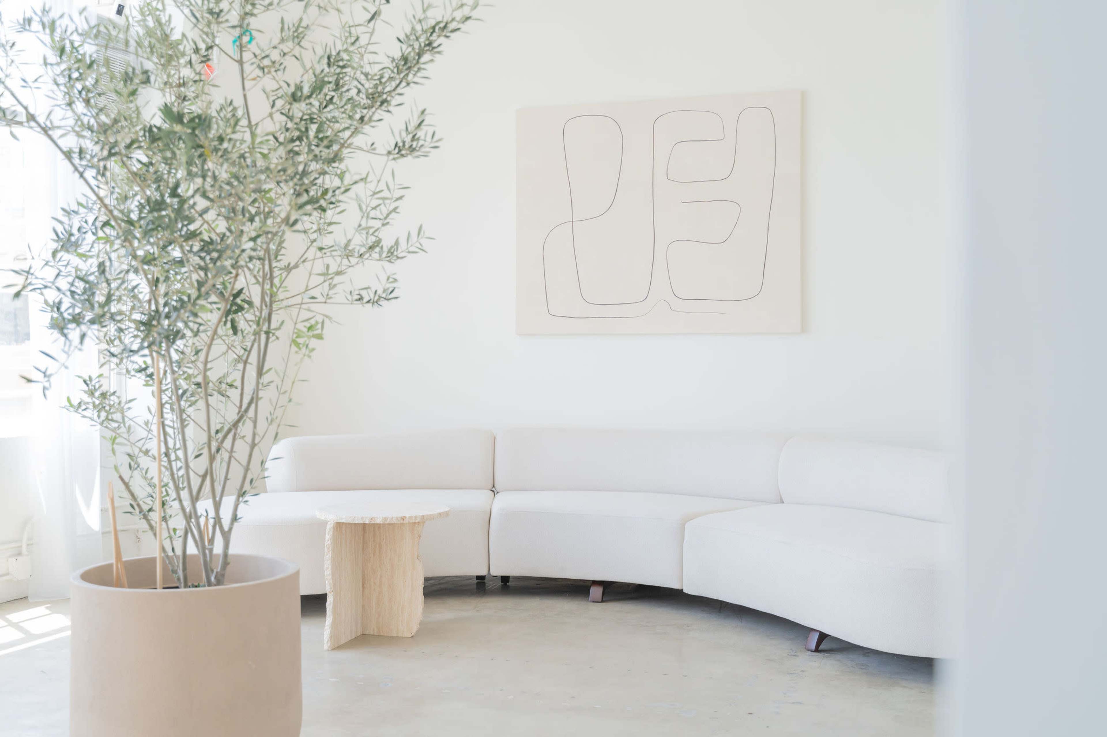 The image shows a minimalist interior space featuring a curved white sofa, a small wooden side table, and a potted plant in a light-filled room with abstract wall art.