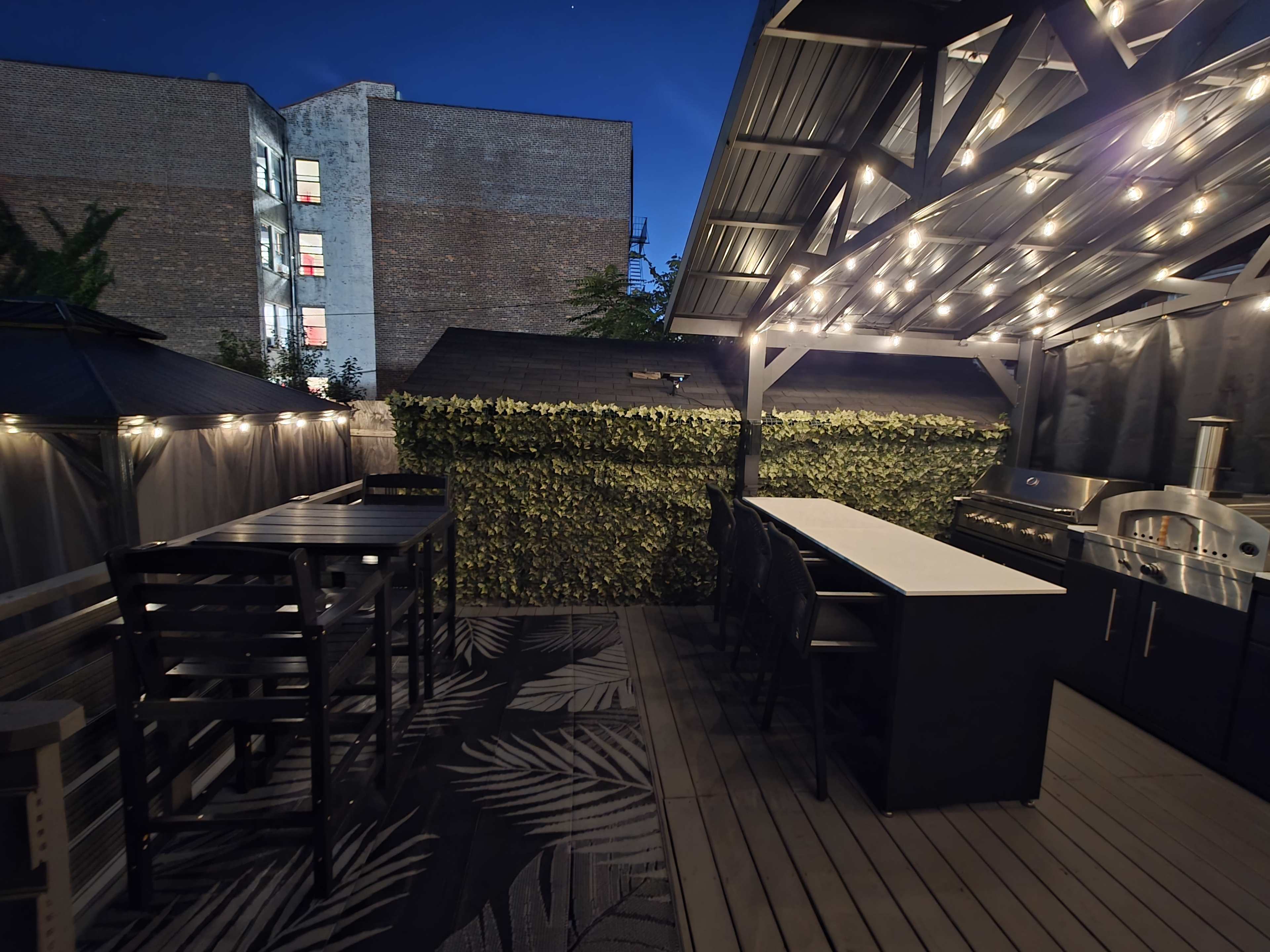 The image shows a rooftop deck with seating areas, a bar counter, and string lights illuminating the space, along with greenery on the walls.
