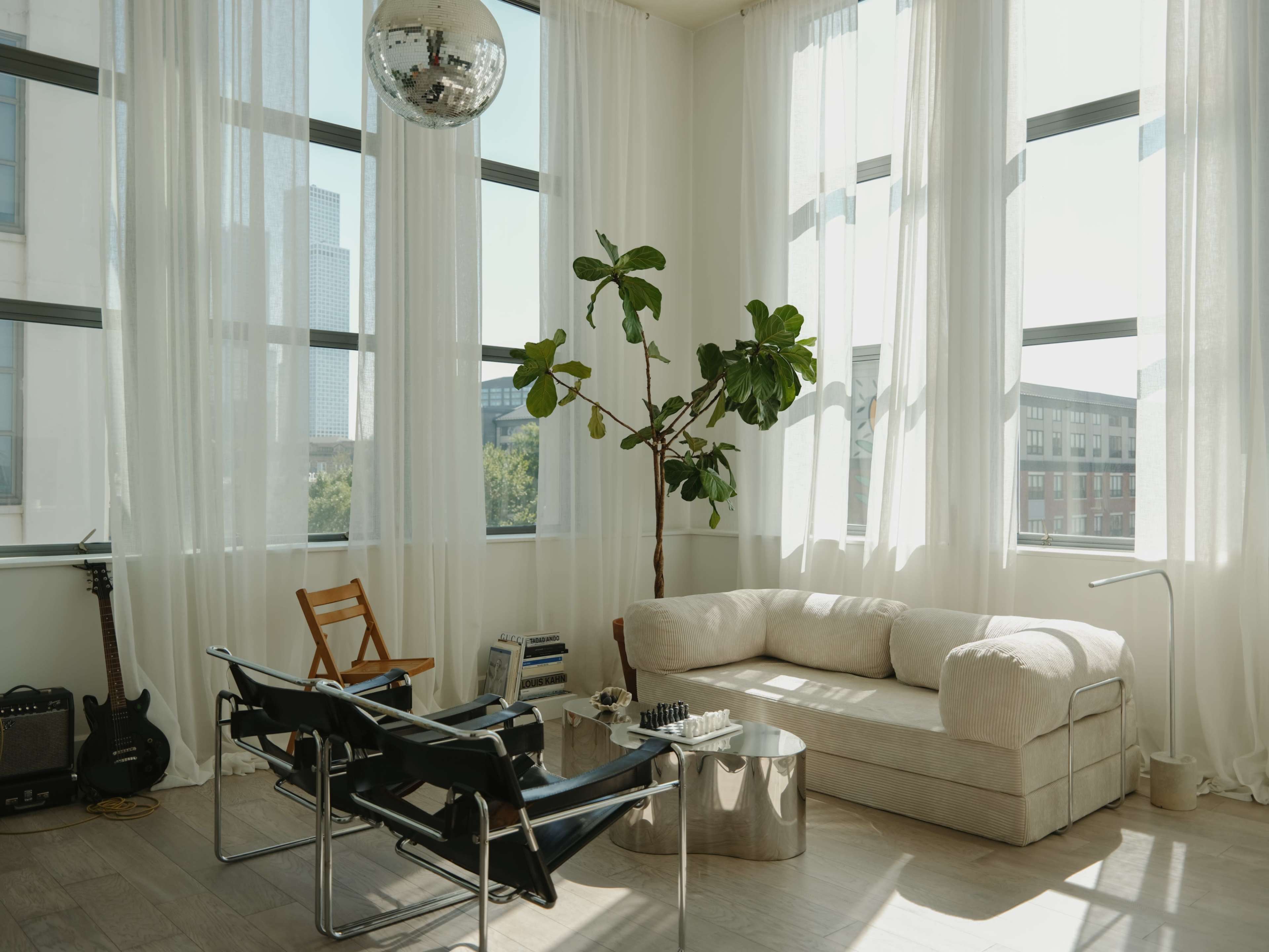 A spacious living room features large windows with sheer curtains, a comfortable sofa, two black chairs, a coffee table, a potted plant, a guitar, and a disco ball hanging from the ceiling.
