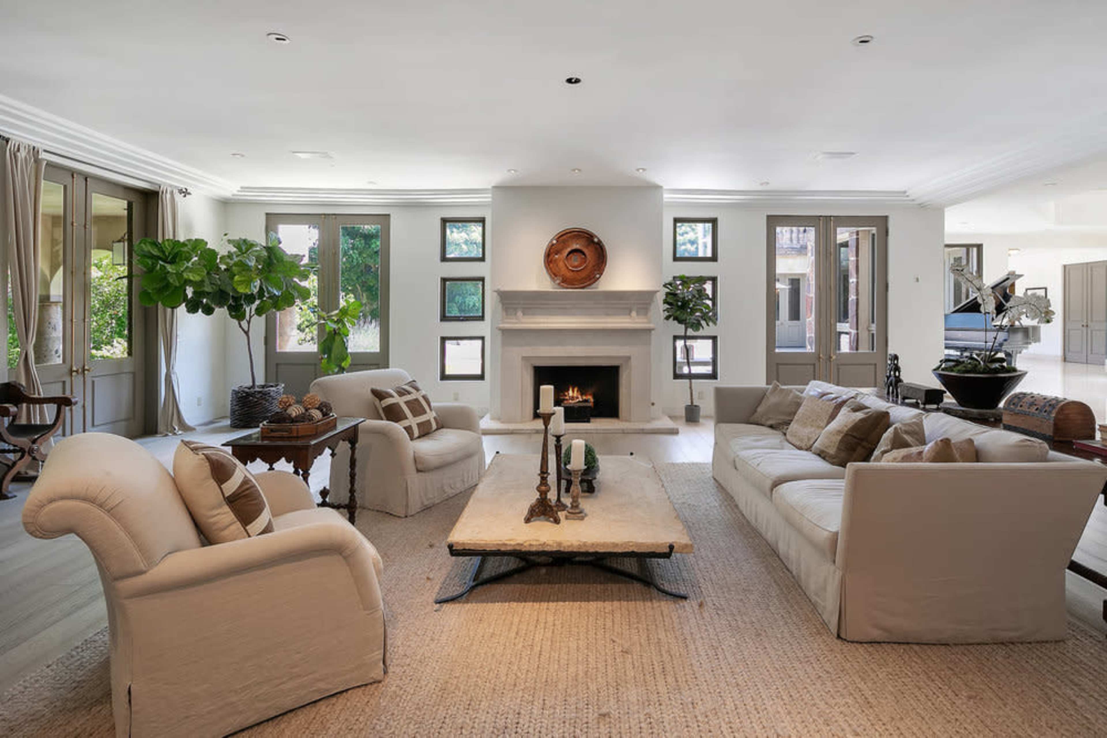 A spacious living room features a large coffee table, two sofas, and a fireplace surrounded by large windows letting in natural light.