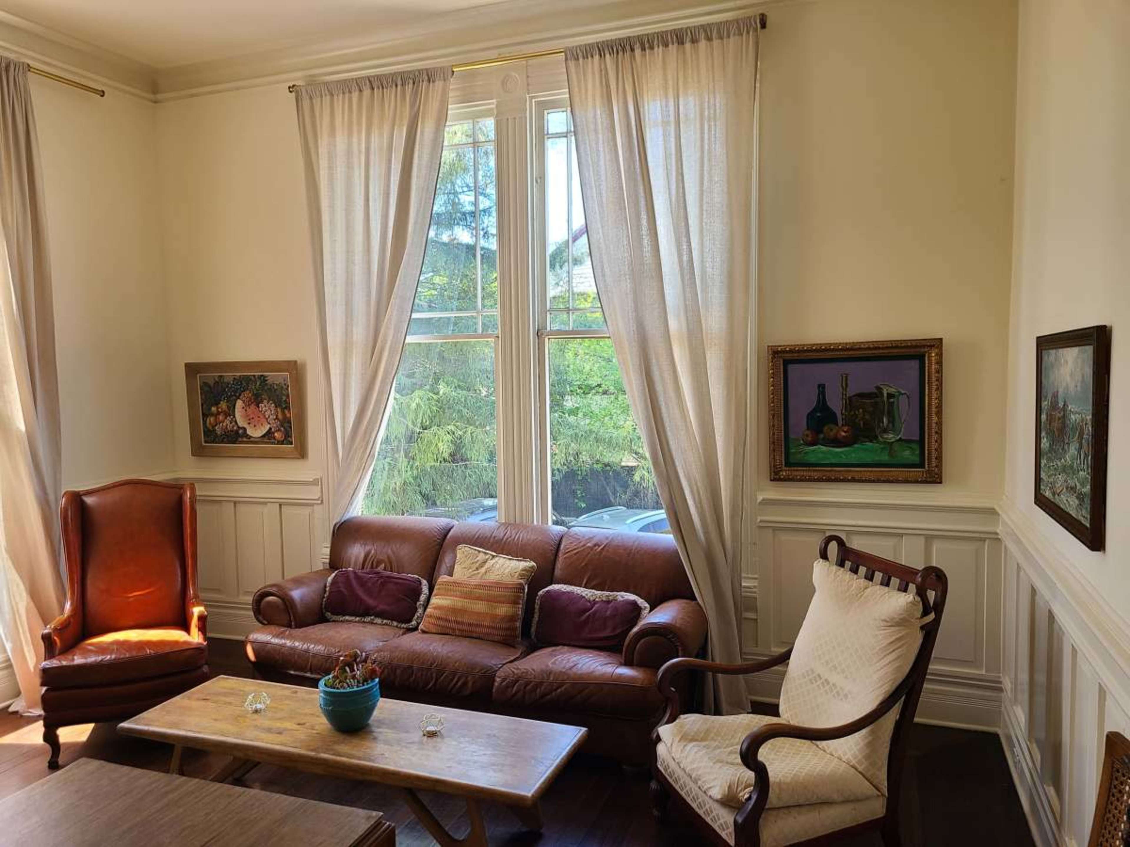 The image shows a cozy living room with a brown leather sofa, two armchairs, a wooden coffee table, and windows covered by sheer curtains, allowing natural light to fill the space.
