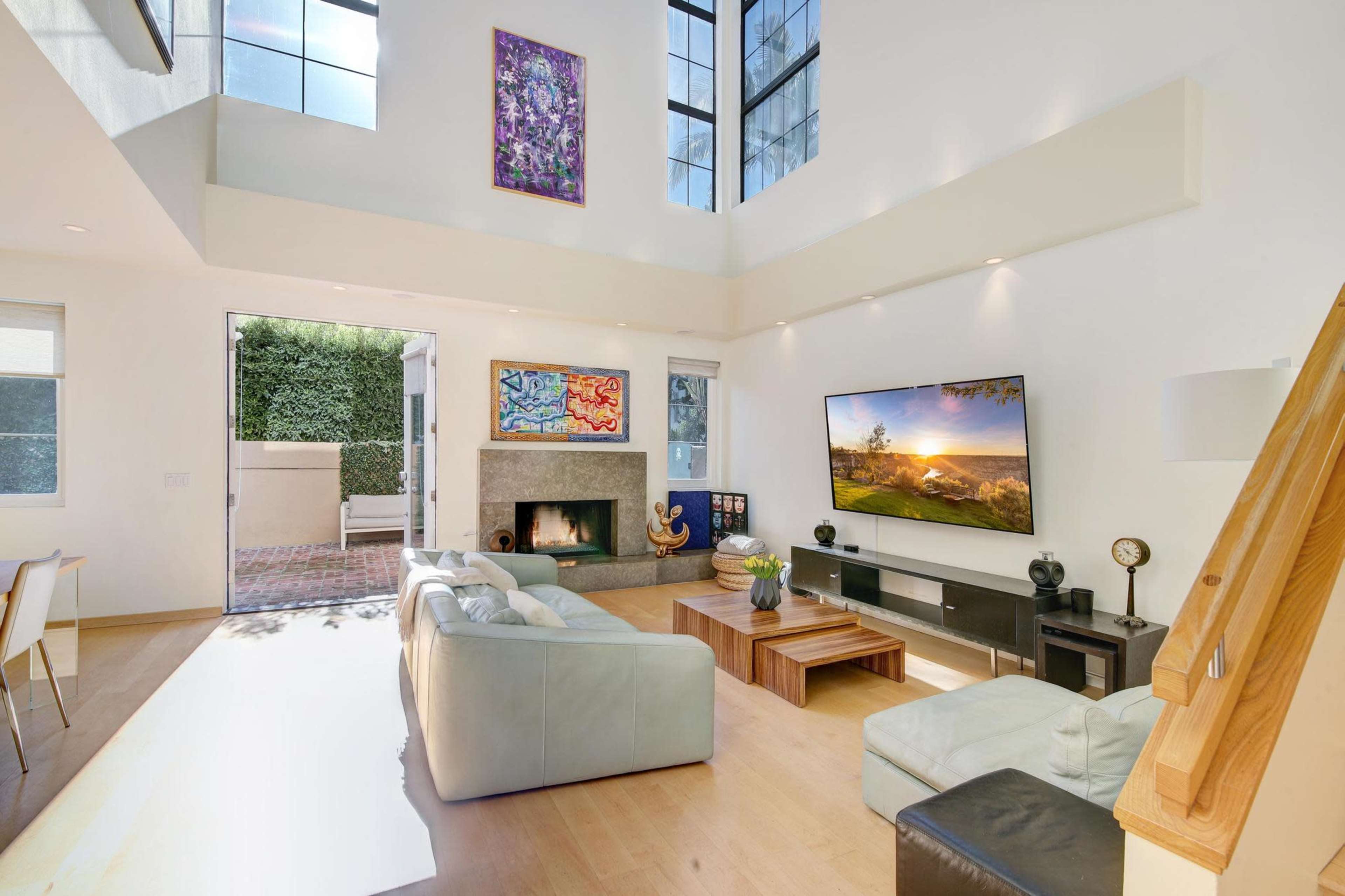 A modern living room with a large TV mounted on the wall, a fireplace, and large windows allowing natural light to enter.