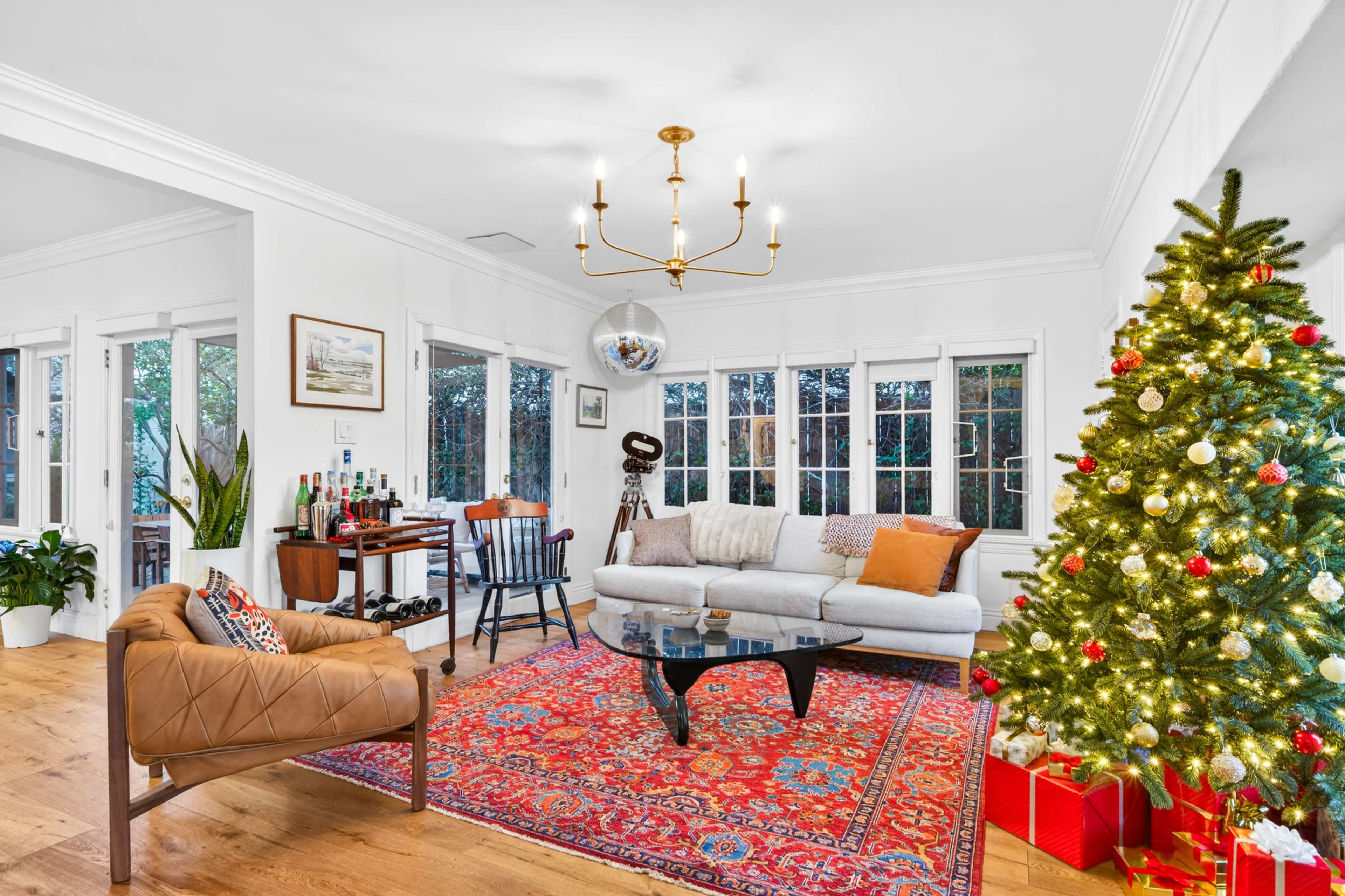 The image shows a stylish living room featuring a Christmas tree decorated with lights and ornaments, a comfortable sofa, a coffee table, and a cozy armchair, with large windows allowing natural light to enter.