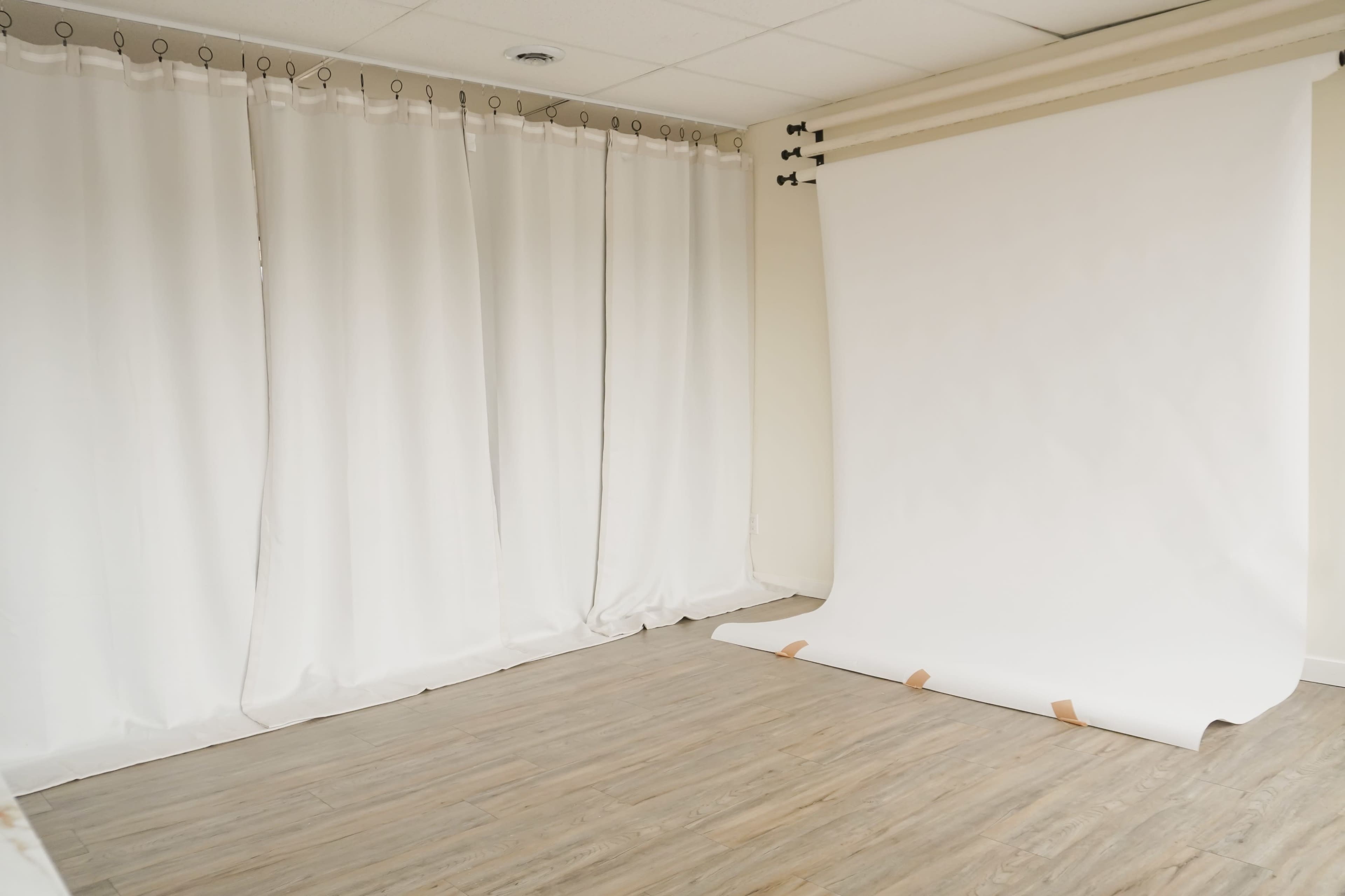 A photography studio with white curtains on one side and a white backdrop leaning against the wall on the other side.