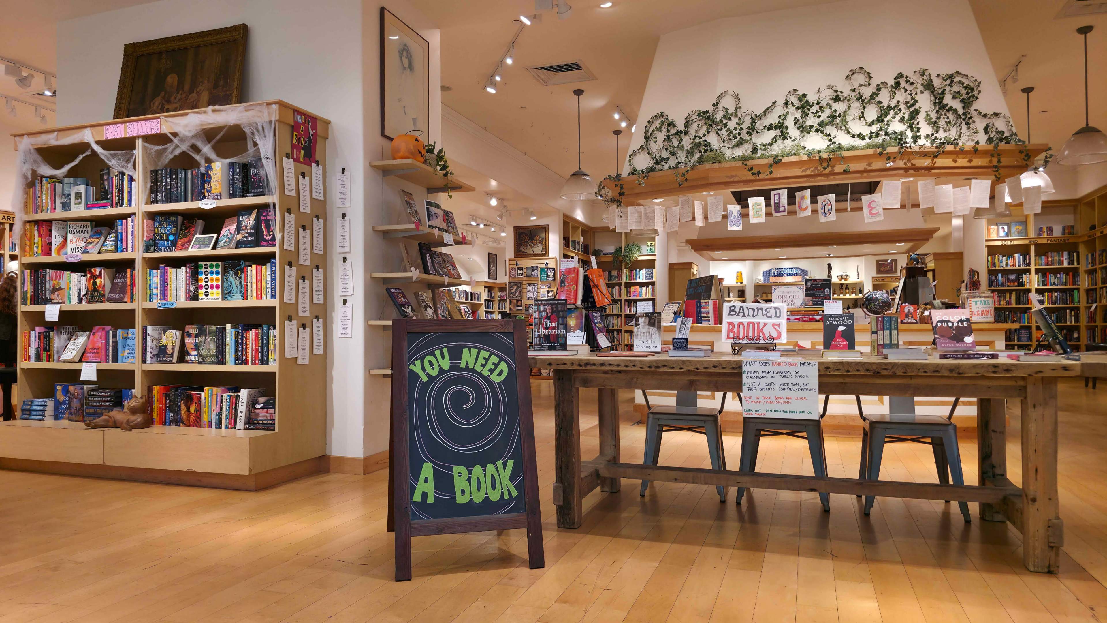 The image shows the interior of a bookstore featuring shelves filled with books, a wooden table with a sign reading "YOU NEED A BOOK," and decorative elements such as vines overhead.