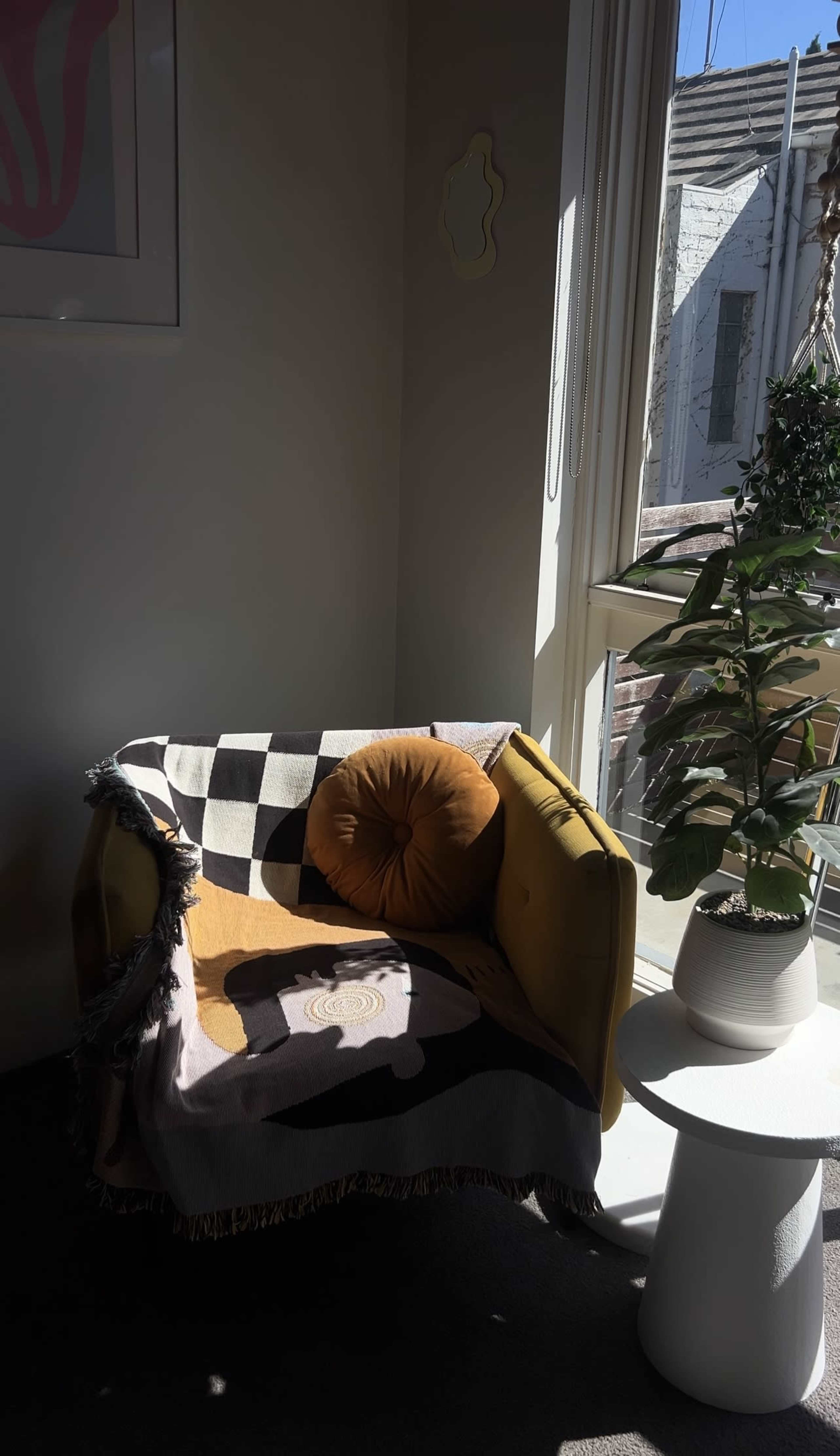 A yellow chair with a patterned blanket and a round cushion is positioned near a window, with a small white table and a potted plant beside it.
