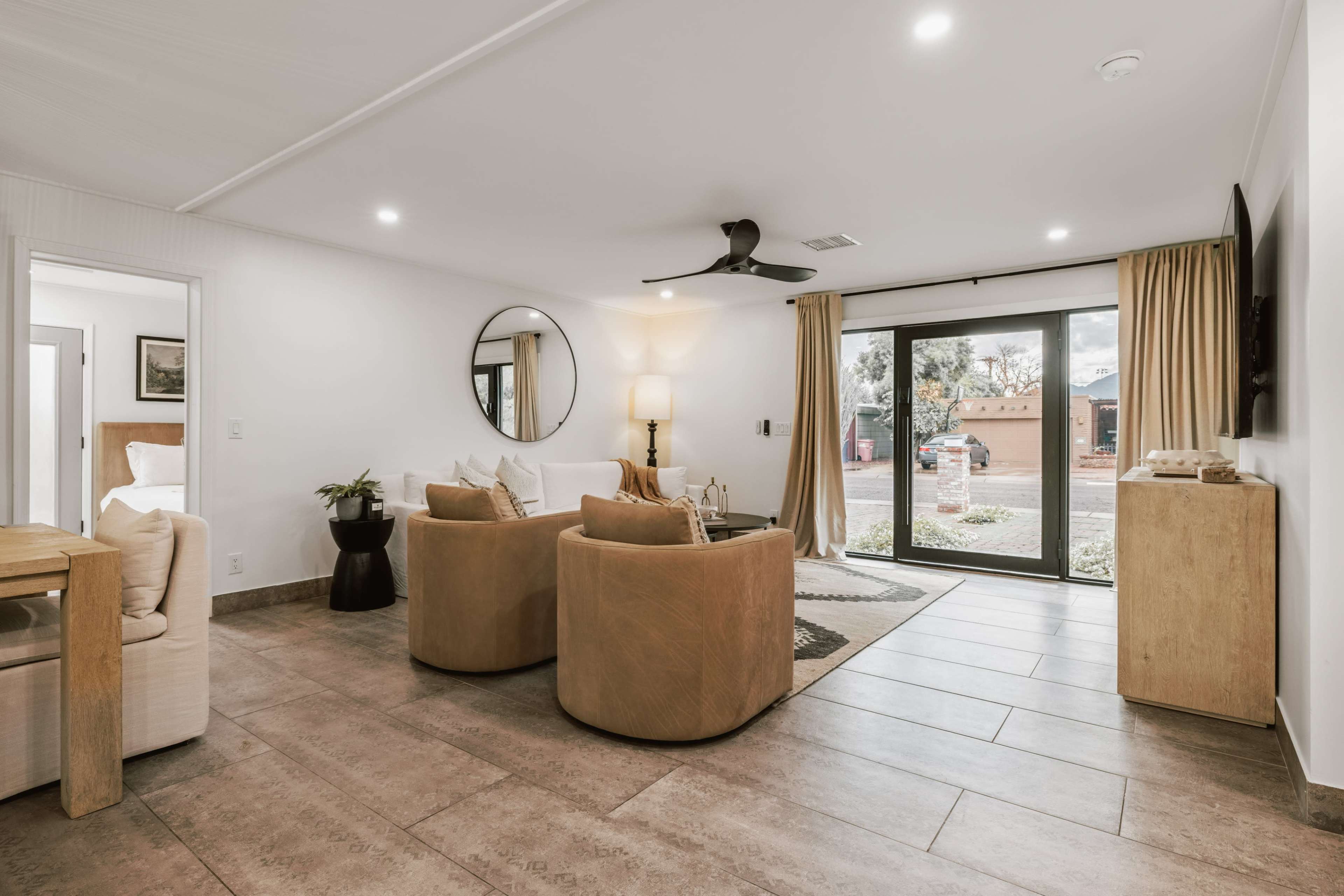 The image shows a modern living room with two round armchairs, a circular mirror, a small table, and sliding glass doors that lead to an outdoor area.