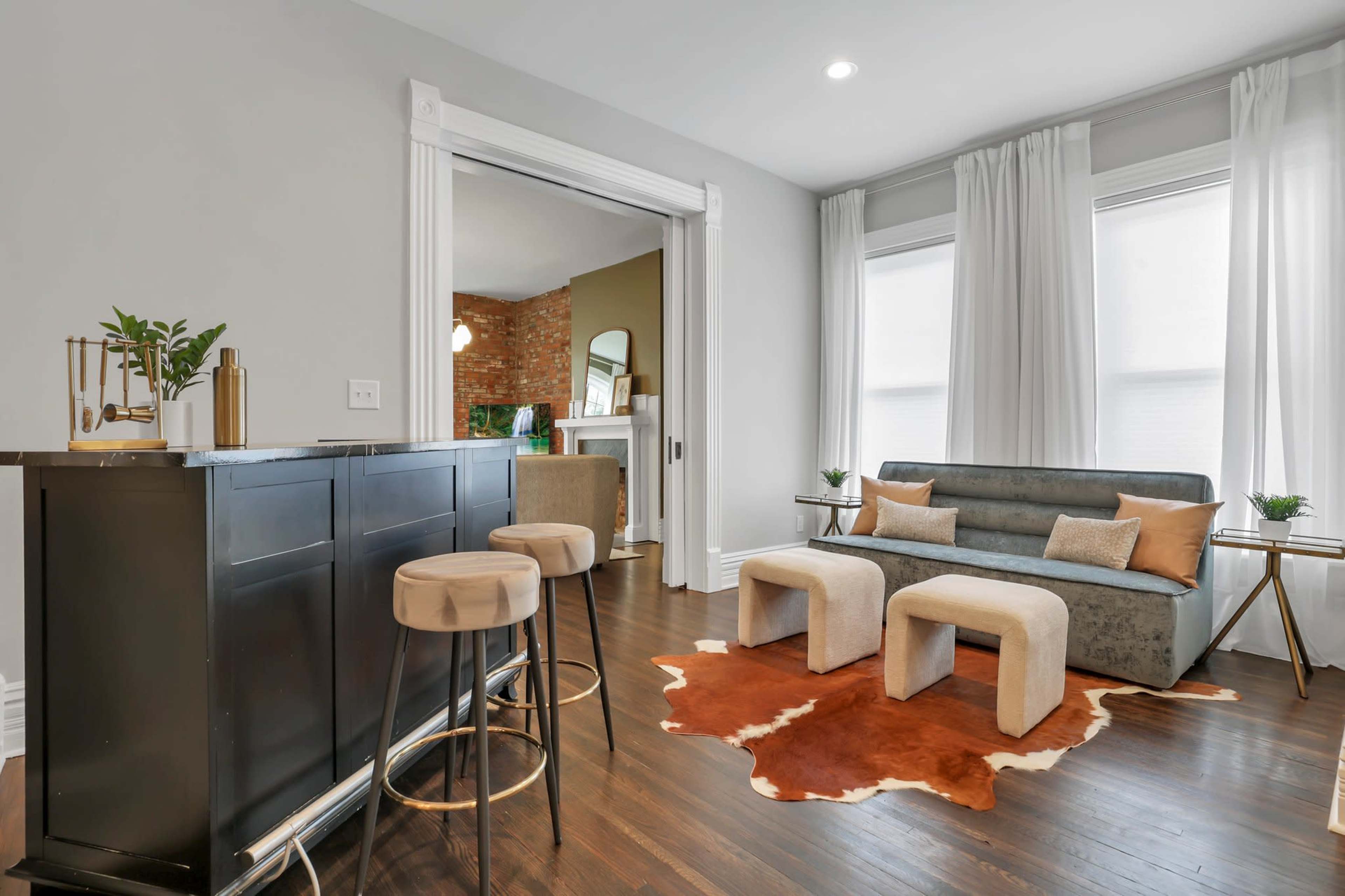 A modern living space with a small bar area, a gray sofa, two light-colored stools, and a cowhide rug on dark hardwood flooring.