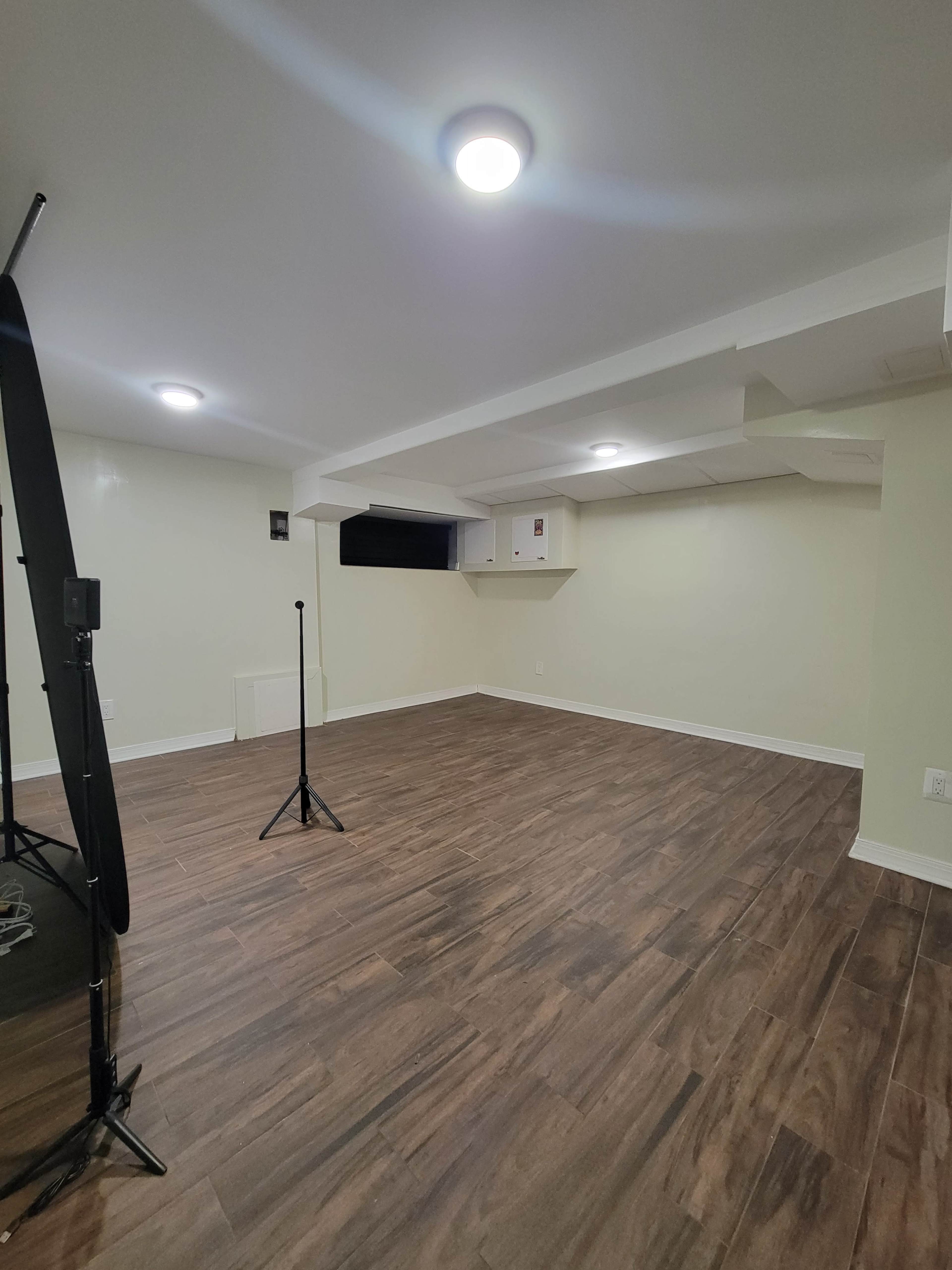 The image shows an empty room with wood-like flooring, white walls, and a low ceiling, featuring two light fixtures and a small window.