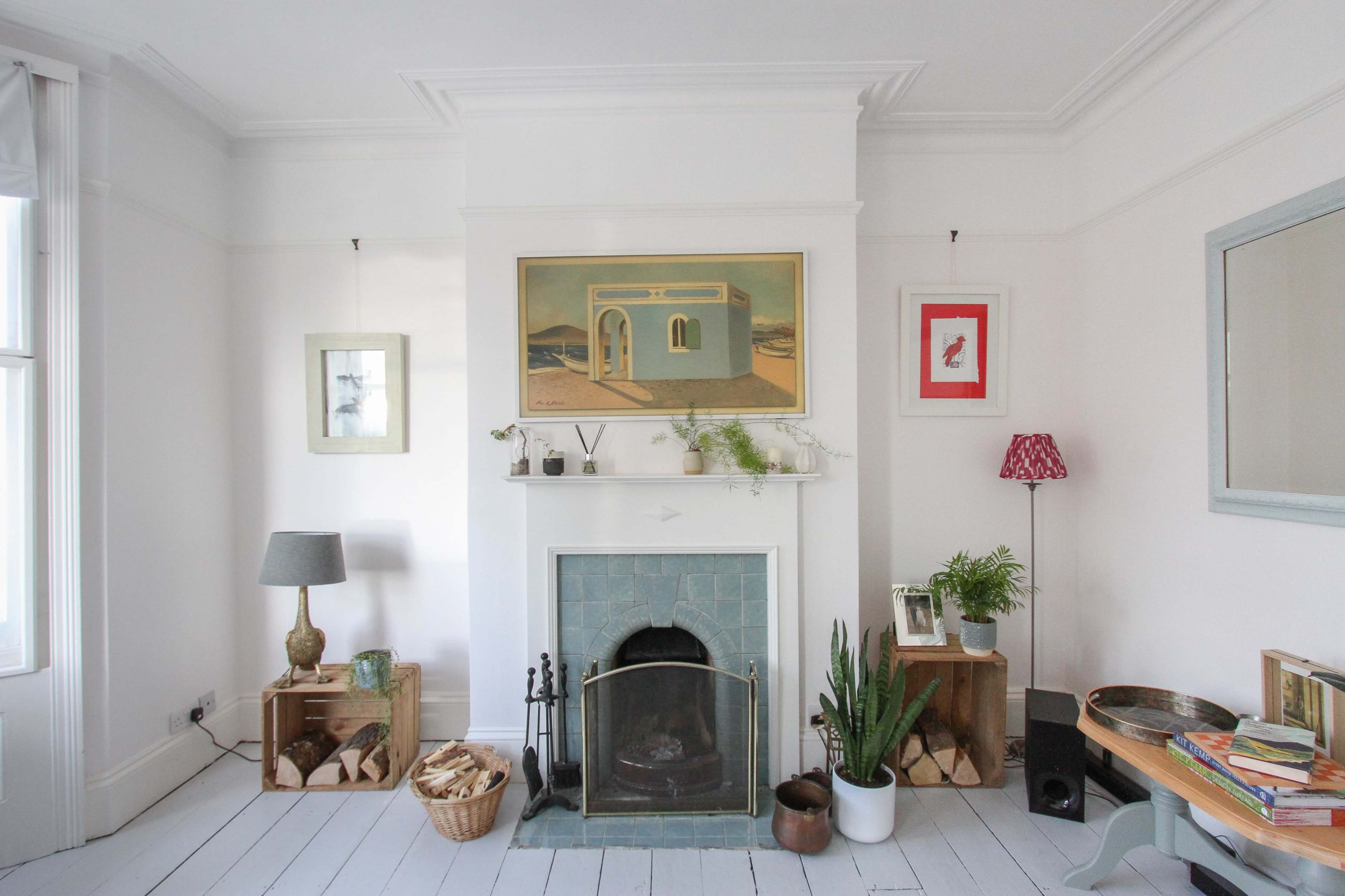 The image depicts a minimalist living room featuring a centered fireplace, two side tables with decorative items, and a mix of plants and art on the walls.