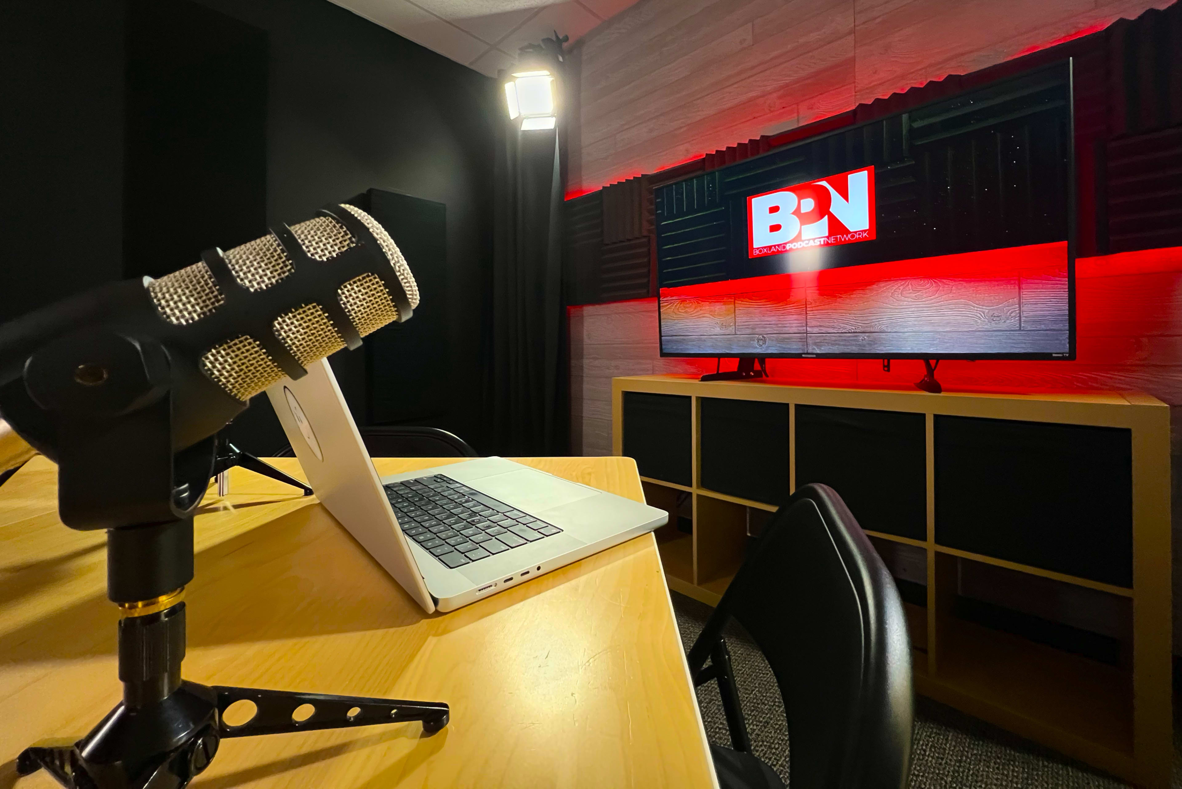 A microphone and a laptop on a wooden desk in a studio setting with a large screen displaying "BN" in the background, surrounded by soundproofing panels.