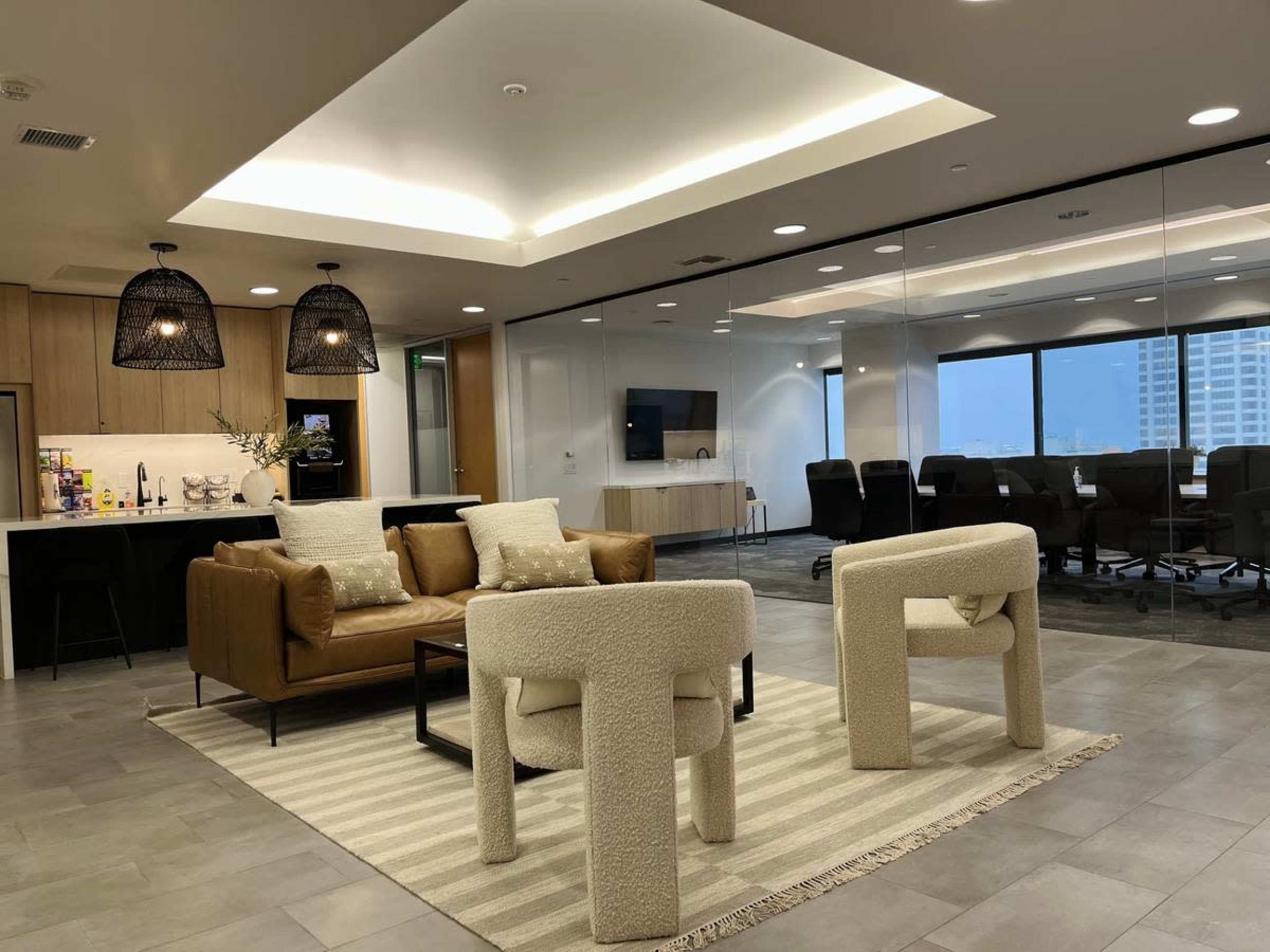 The image shows a modern office lounge area with a brown leather sofa, two upholstered chairs, a large rug, and a glass-walled conference room in the background.