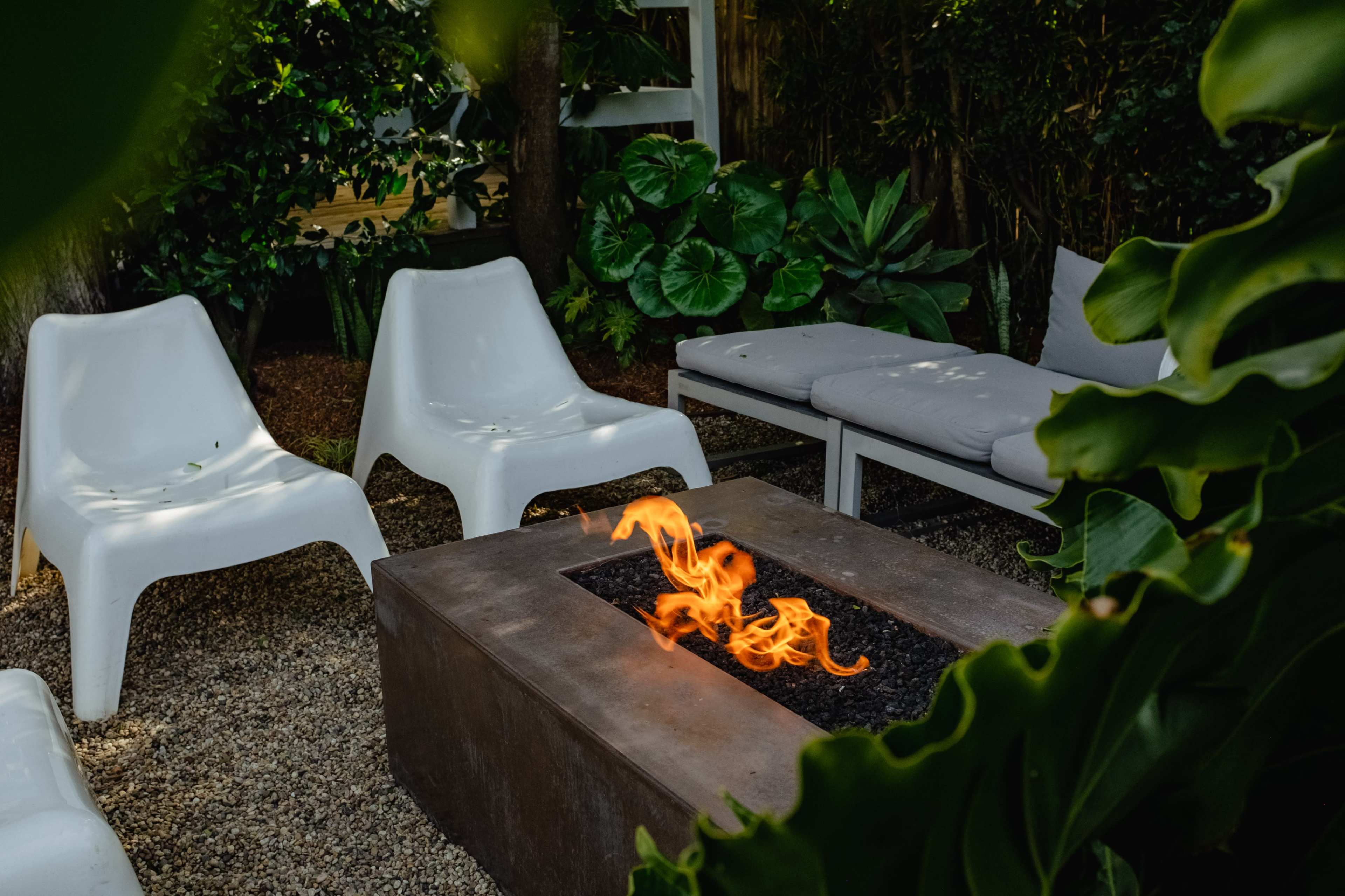 A fire pit is surrounded by white chairs and a lounge area, set among lush green plants in a garden.