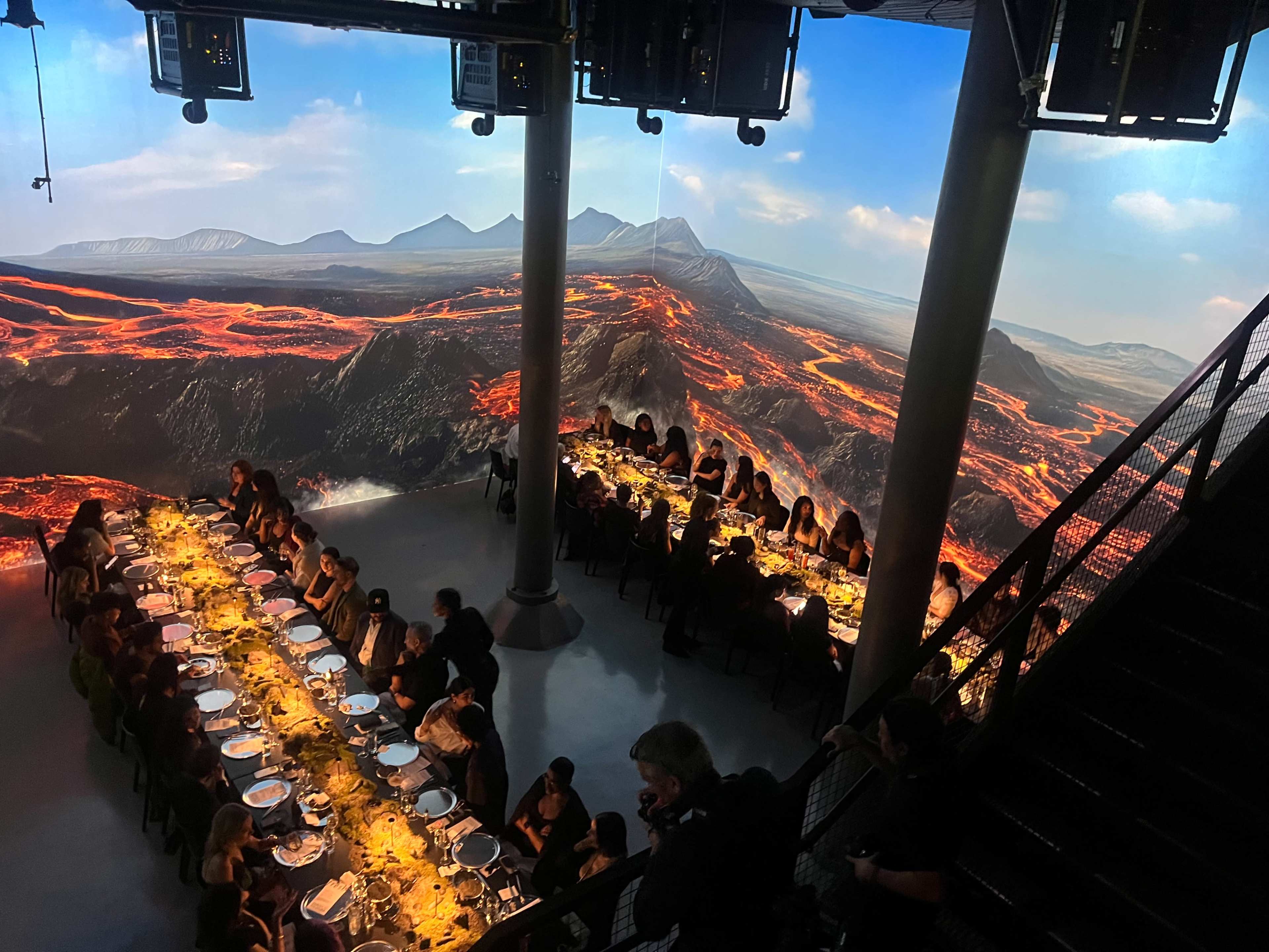 A long dining table filled with guests is set against a backdrop of a vibrant, projected volcanic landscape.
