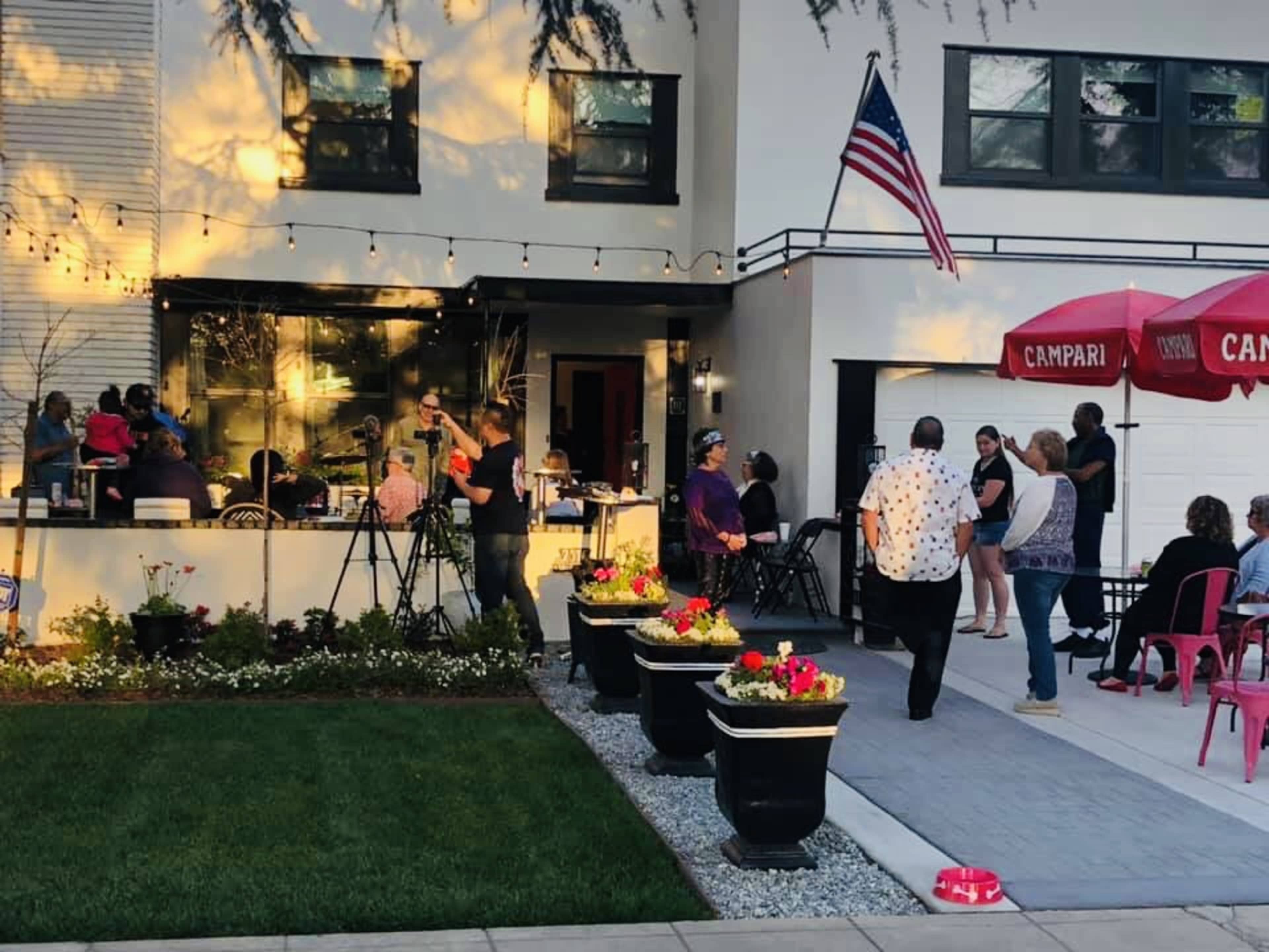 A gathering is taking place outside a modern house, with people mingling and cameras set up around a patio area.