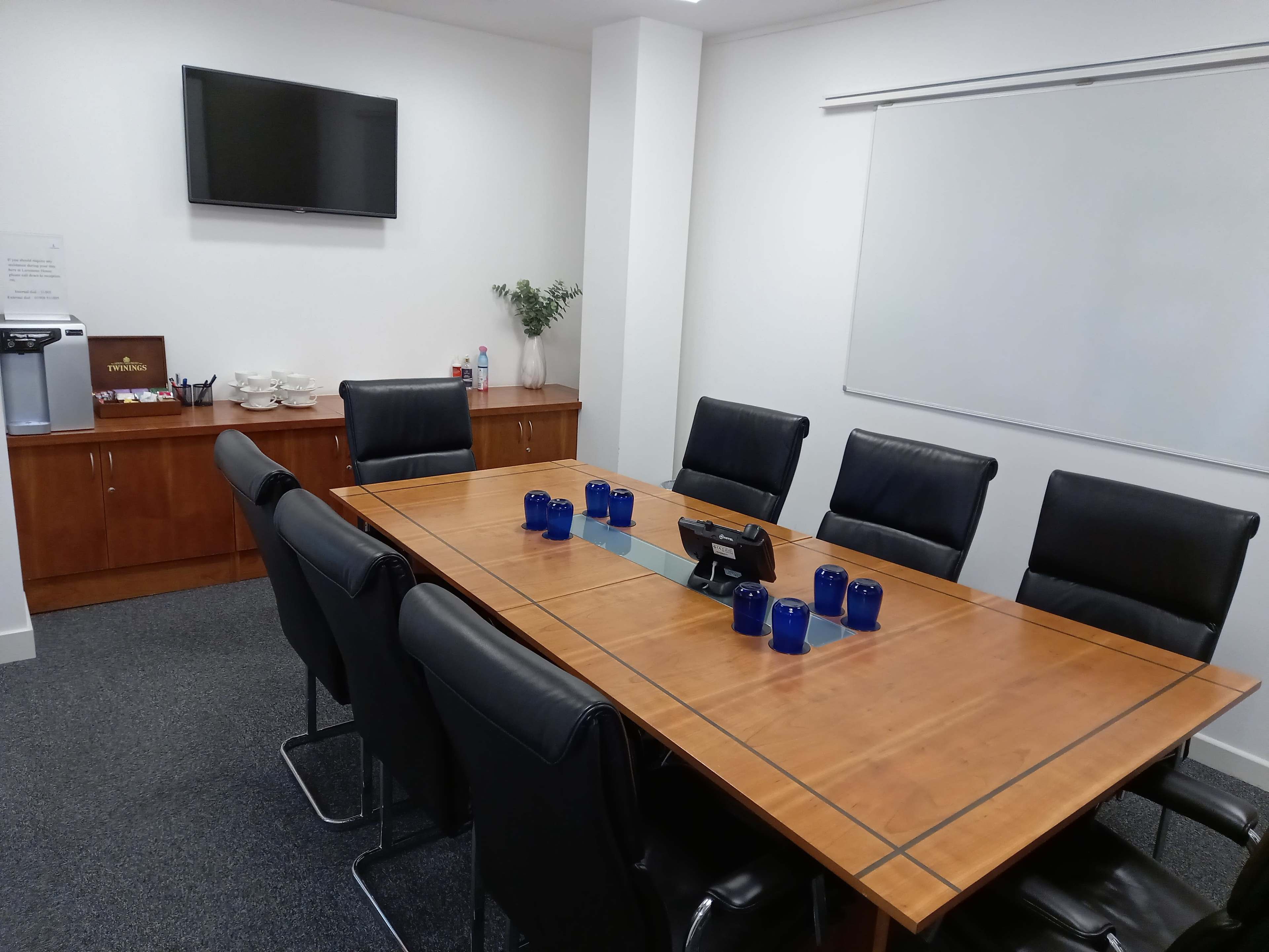 A conference room featuring a large wooden table surrounded by black leather chairs, with a television mounted on the wall and a whiteboard nearby.