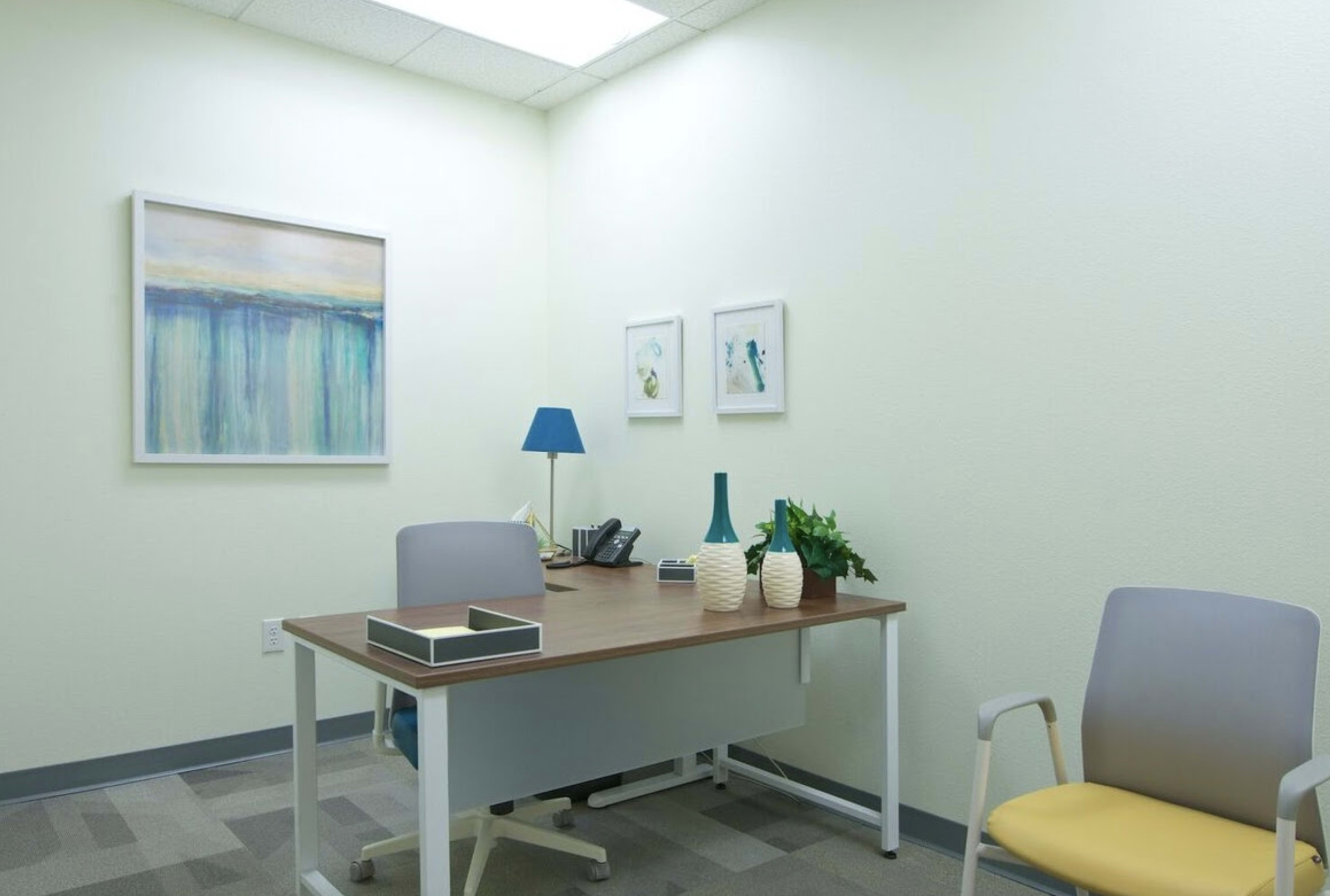 A small office space with a wooden desk, two chairs, a telephone, and decorative items including a lamp and artwork on the walls.