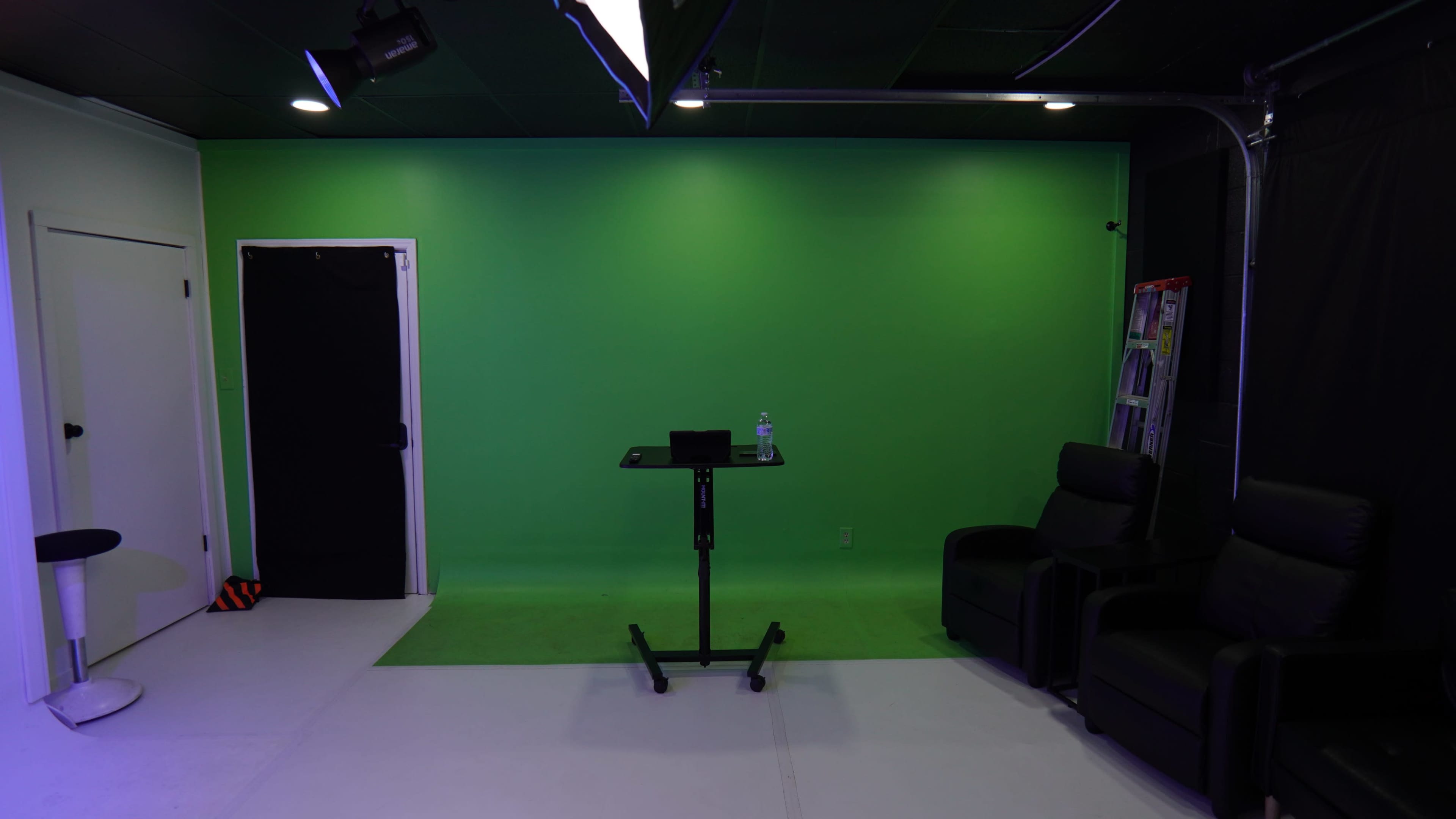 The image shows a green screen studio with a black door on the left, a table with a water bottle, and two black lounge chairs.