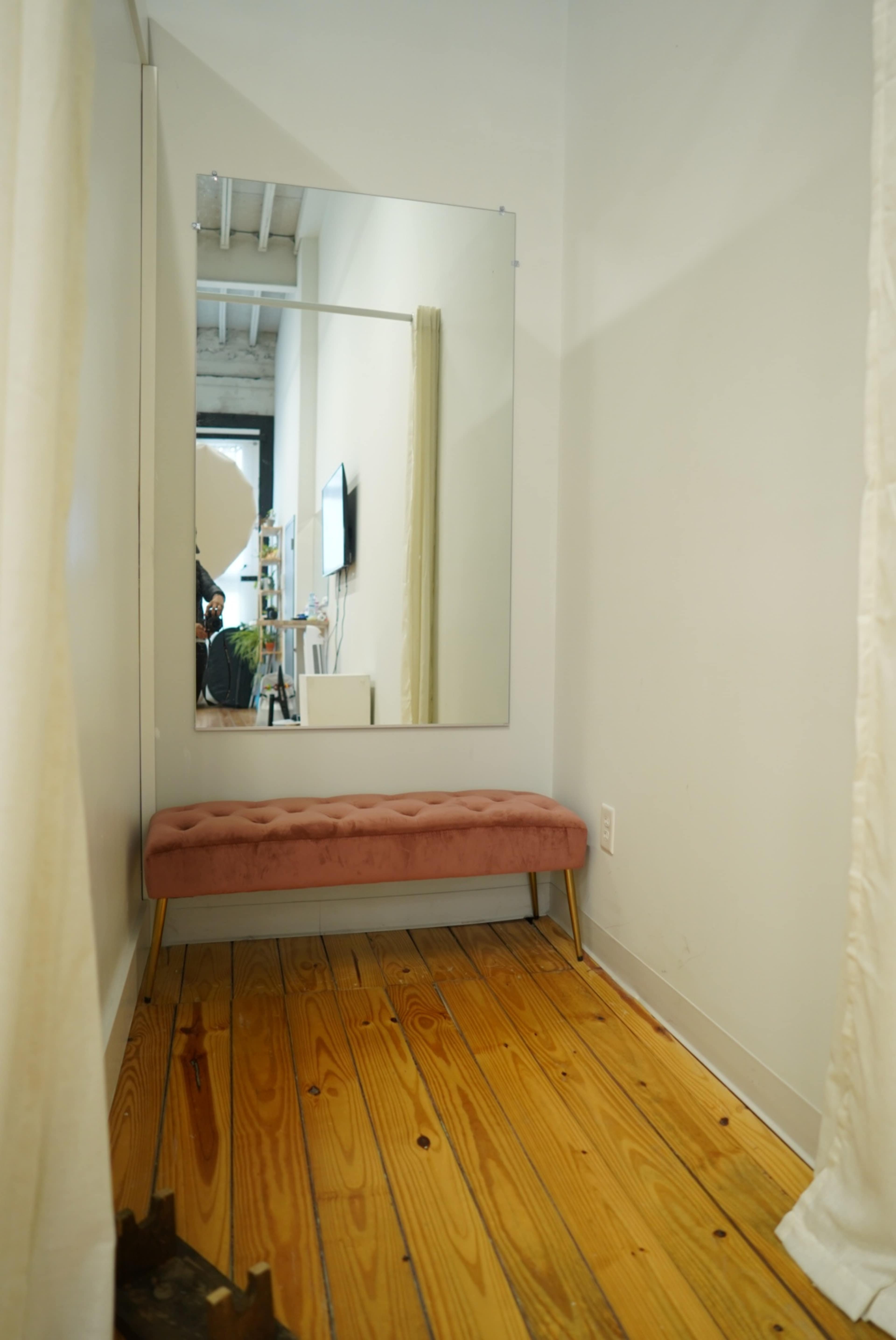 The image shows a narrow hallway with a pink tufted bench and a large mirror on one wall, flanked by a curtain.