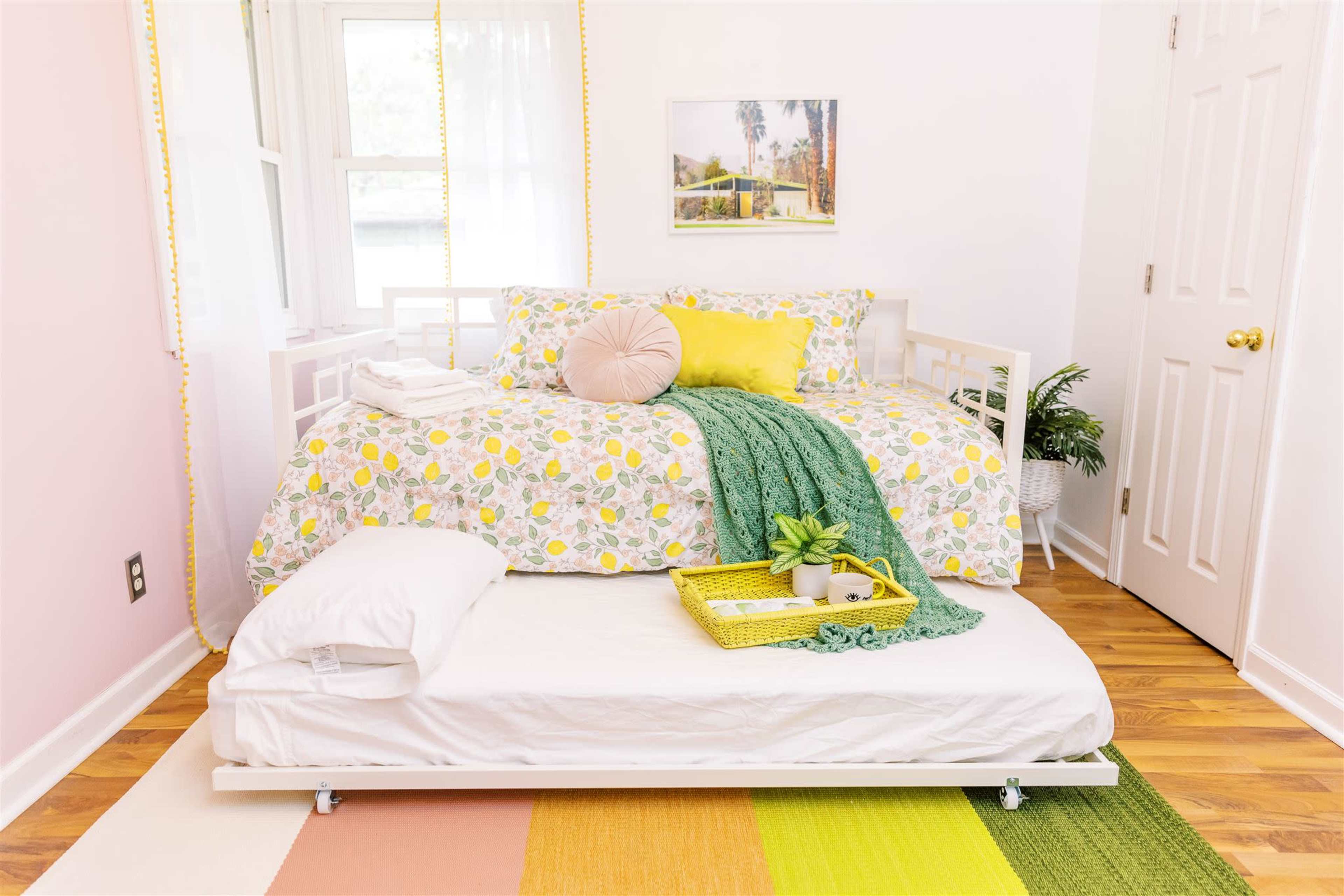 A bright and neatly arranged bedroom featuring a bed with a colorful duvet, a pull-out trundle bed underneath, and decorative elements like a green throw and a tray.